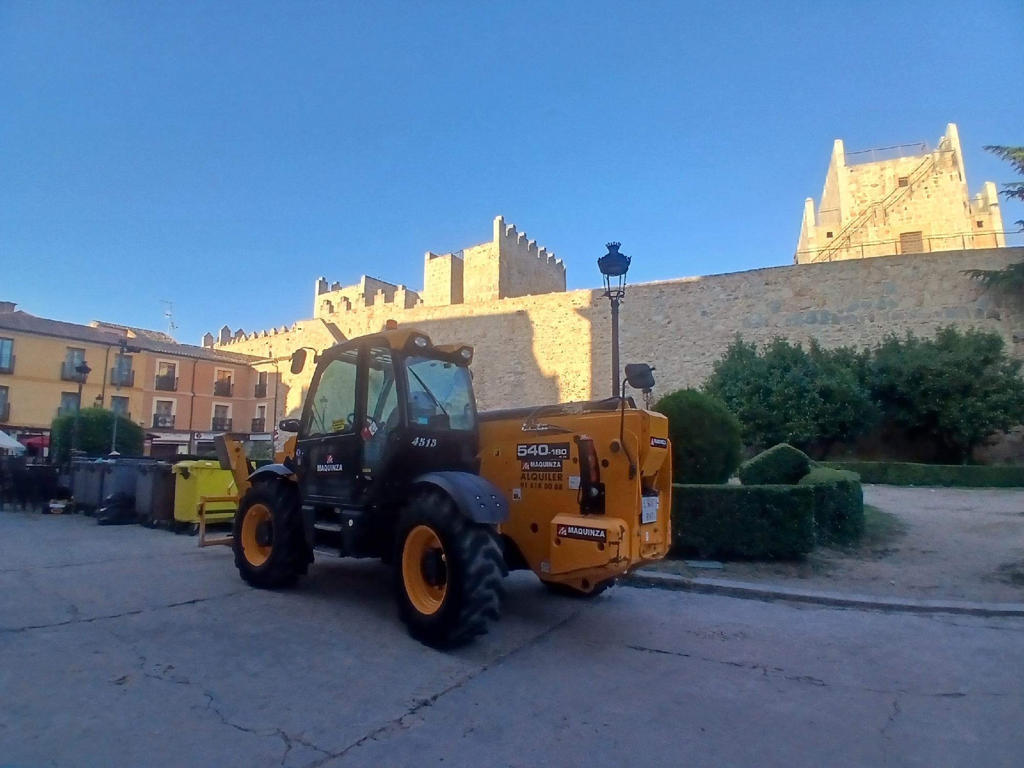 A telehandler, identified by the "MAQUINZA" logo and model number "540-180", is parked on a paved surface in front of a stone wall. The telehandler is yellow and black. Behind the telehandler and wall, buildings with light-colored facades and windows are visible. The sky is clear and blue. The scene appears to be in an urban environment, possibly a public square or street in Ávila, Spain, given the architecture of the buildings and the style of the wall, which resembles the ancient city walls of Ávila. There are no people visible in the image. Several large trash bins are also present to the left of the telehandler.