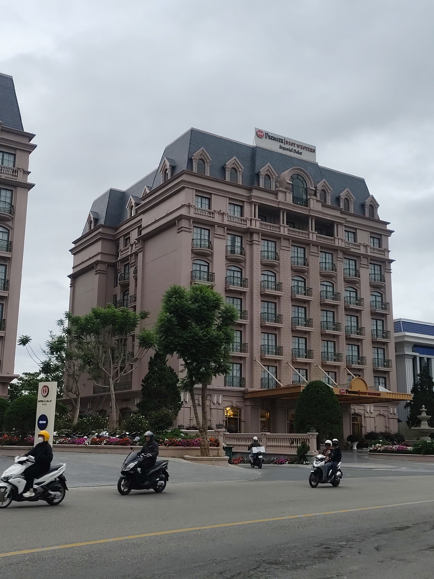 The image captures a street scene in Lâm Viên - Đà Lạt, Vietnam, featuring the Premier Best Western Imperial Dalat Hotel. The hotel, a multi-story building with a light pink facade and dark grey mansard roof, dominates the background. It has numerous windows, each with a small balcony. A sign bearing the hotel's name is visible on the roof. At street level, several scooters with riders are present, moving along a road marked with yellow lines. The foreground is adorned with flowerbeds. The sky is overcast, casting a diffused light over the scene.