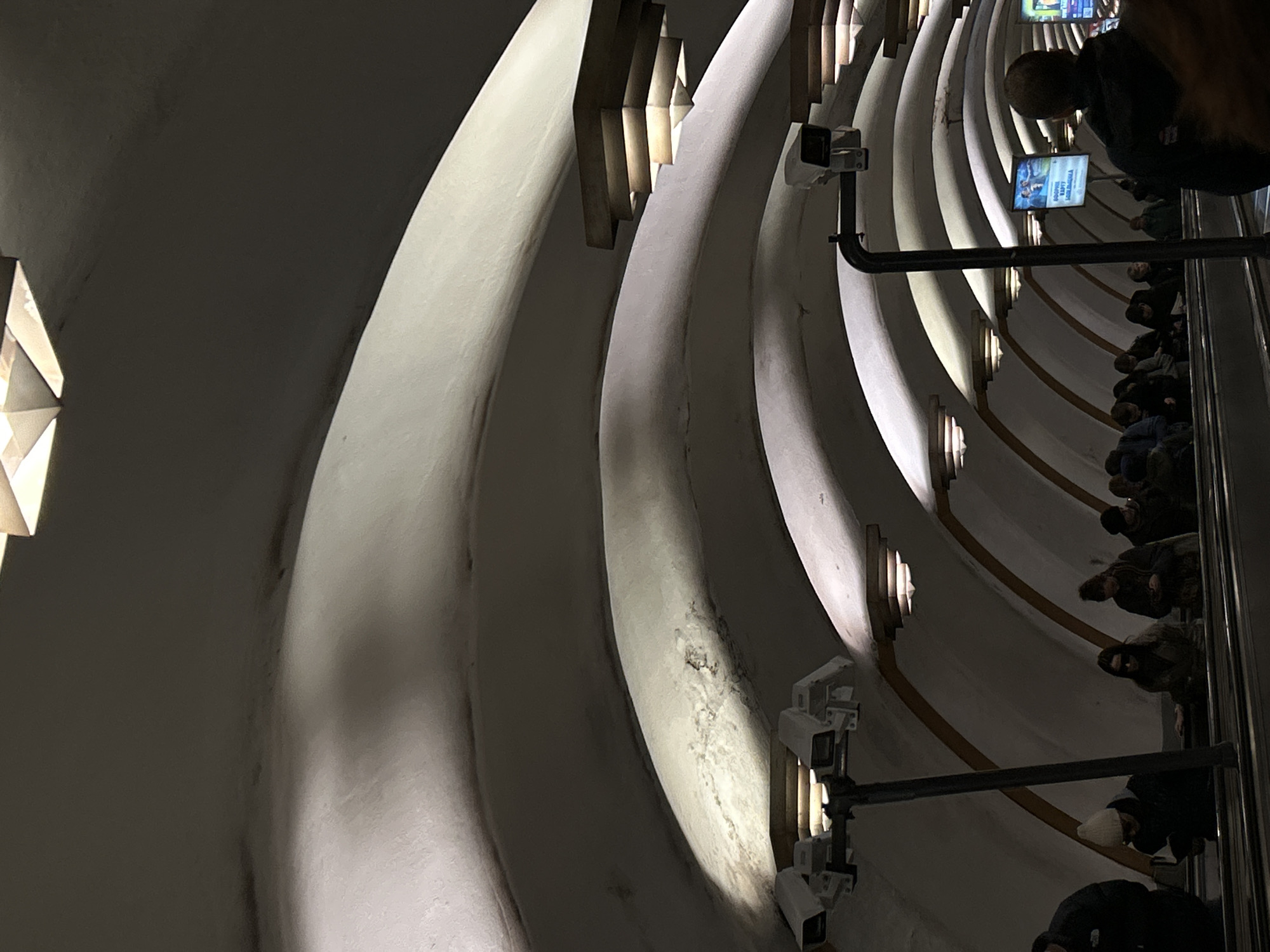 This image captures a view inside a deep, spiraling underground passage, characteristic of a subway station, likely in Saint Petersburg, Russia. The dominant features are the curved, light-colored walls, segmented into distinct ribbed sections that form a continuous helix. These walls are illuminated by integrated lighting fixtures, which appear as rectangular or diamond-shaped elements casting strong shadows and accentuating the architectural curves.
On the right side of the image, an escalator or moving walkway is visible, carrying a crowd of people. Most individuals are seen from behind or from above their heads, dressed in dark clothing, including jackets and hats, suggesting cool weather. A dark metal handrail runs alongside the escalator.
Multiple white security cameras are mounted on the walls, observing the flow of people and the passage. Higher up on the right, two illuminated rectangular screens display advertisements with Russian text. Visible words include "БОНУСЫ" (Bonuses) and "КАРТУ" (Card), indicating a promotional message.
The scene depicts a busy public transport environment, with artificial lighting creating a sense of depth and scale within the subterranean space. The exact time of day is not discernible due to the underground setting.