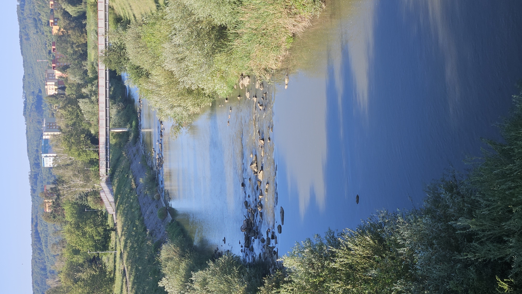The image captures a serene view of the Sekčov River flowing through the city of Prešov, Slovakia. The river, bordered by lush green banks and vegetation, meanders through the urban landscape. In the background, residential buildings and trees dot the hillside, suggesting a harmonious blend of nature and city life. A pedestrian bridge spans across the river, providing a scenic pathway for onlookers. The water reflects the clear blue sky and the surrounding greenery, creating a tranquil atmosphere. The scene is bathed in the warm light of the sun, casting gentle shadows on the water's surface. The presence of rocks and shallow areas in the river adds to its natural charm.