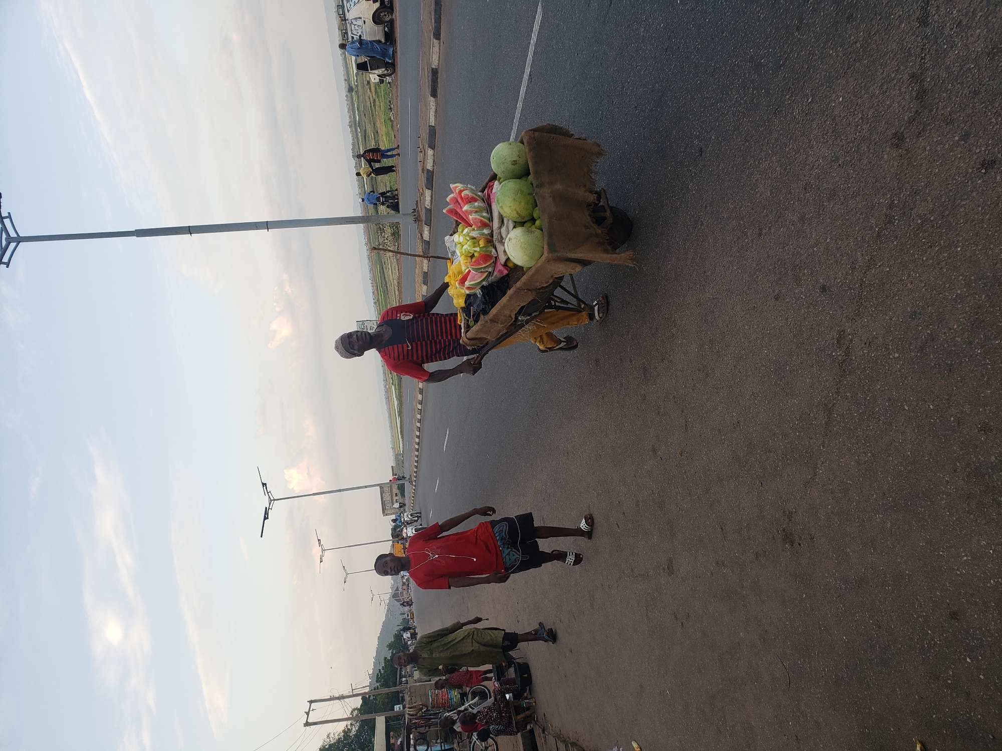 This outdoor photograph, taken in Lokoja, Nigeria, captures a bustling roadside scene under a bright, slightly cloudy sky. The image features a primary subject, a street vendor, centrally positioned as he pushes a fruit-laden cart along the asphalt road.
The vendor, a man wearing a red and black striped shirt, yellowish-brown trousers, and a dark cap, is actively pushing a metal-framed pushcart. The cart is covered with brown burlap and filled with a variety of fresh produce. Prominently displayed are several whole, round green melons (likely watermelons) and numerous vibrant red watermelon slices, individually packaged in clear plastic bags. Other smaller green fruits are also visible in bags.
The setting is a paved asphalt road, likely a highway or main thoroughfare, indicated by a faint white lane divider running down its length. Several other people are present on or near the road. Closer to the foreground, a man in a red t-shirt, patterned dark shorts, and sandals stands with earphones in. Slightly behind him, another person in a green long-sleeved garment stands next to what appears to be a small makeshift stall or collection of items. In the distance, multiple individuals, cars, and motorbikes can be seen along the roadside.
Tall metal streetlights with modern fixtures line the road. Beyond the immediate roadside, a flat, open landscape stretches to the horizon, where distant hills or mountains are faintly visible under the soft, diffused daylight. The weather appears clear with light, wispy clouds, suggesting either an early morning or late afternoon light. The photograph captures a typical scene of roadside commerce and pedestrian activity in the region. No visible text is discernible in the image.