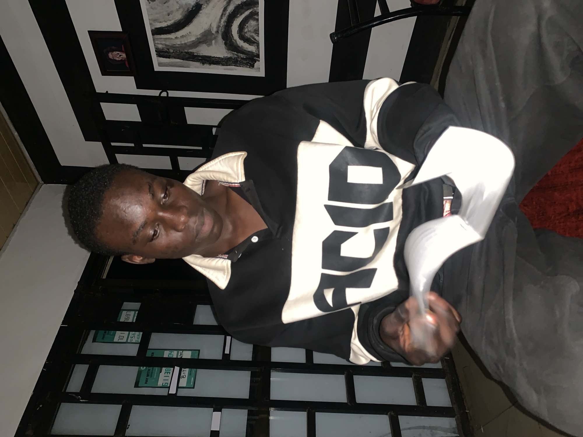 A young adult male, of apparent Black ethnicity, is seated indoors in Jos, Nigeria, in dim artificial lighting. He is wearing a black, white, and cream long-sleeved polo shirt with the word "ACID" prominently displayed across the chest, and dark trousers. His gaze is directed downwards, focused intently on several white papers he holds open in his hands, appearing to be reading or reviewing them. Minor blemishes are visible on his forehead.
To his left, behind him, a window with dark metal security bars is visible. Affixed to the window are multiple green rectangular signs, partially legible, indicating payment options such as "PAY WITH TRANSFER" and "BITCOIN," along with various numbers like "701," "056," and "61138."
To his right, the wall features a distinct black and white horizontal stripe pattern. A small framed photograph depicting a person with an open-mouthed expression is mounted higher on the wall, above a larger framed piece of abstract art with black and white swirling patterns. A dark chair back and leg are partially visible on the far right. A reddish textile, likely a cushion or rug, is present on the floor in the lower right foreground.