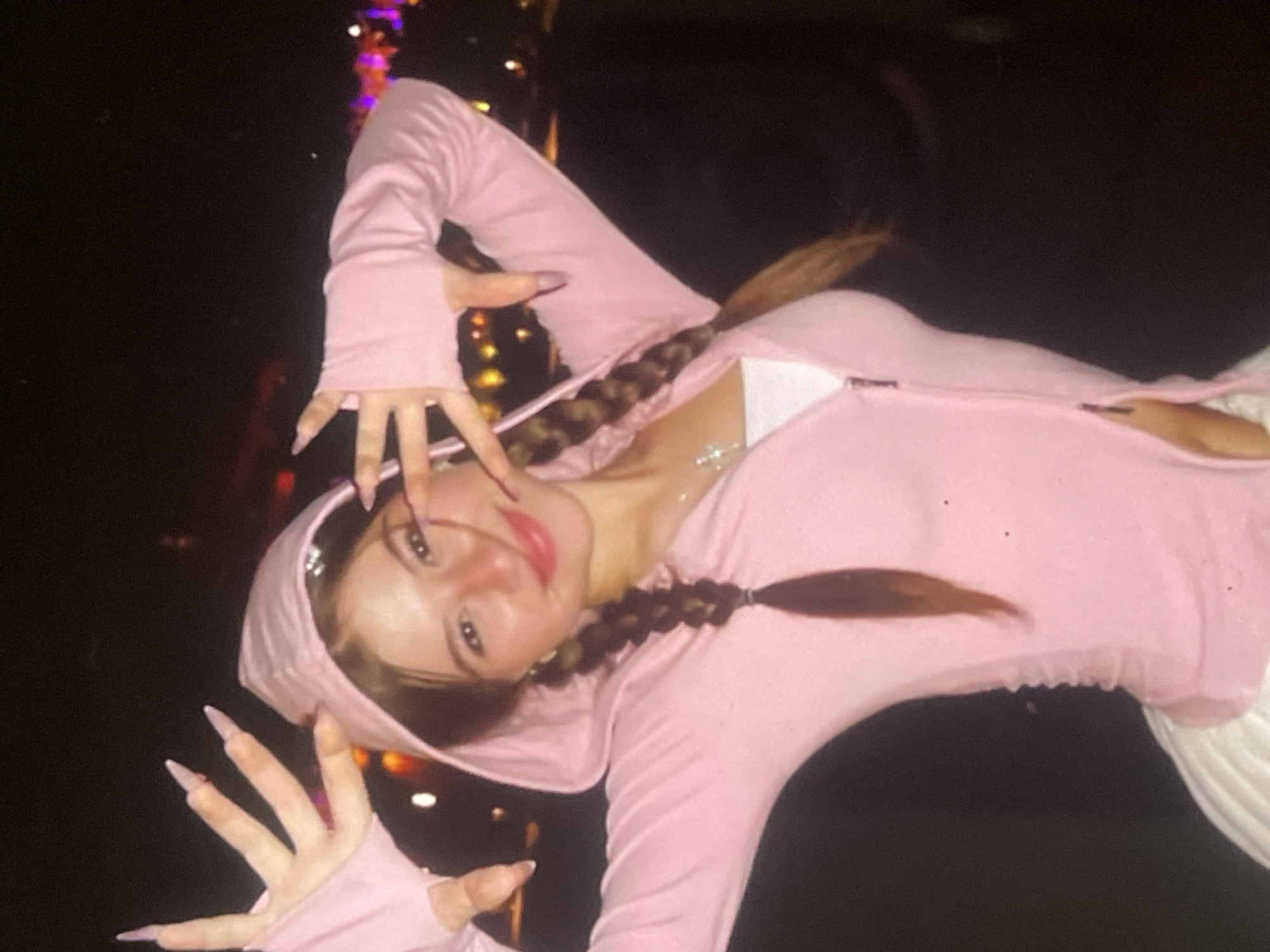 A young woman is captured in a dynamic pose, set against a dark, blurred background with scattered, out-of-focus lights, suggesting a nighttime scene in Poltava, Ukraine, possibly at an event or entertainment venue.
The subject is a young woman with dark hair styled into two long braids, one resting on each side of her chest. She is wearing a light pink, long-sleeved hooded top or jacket, partially unzipped to reveal a white garment underneath. The sleeves extend over her hands, leaving her fingers exposed, which display long, pointed artificial nails in a light pink or purple shade. A subtle, sparkling necklace is visible around her neck. She appears to be wearing white trousers or a skirt on her lower half.
She is looking directly at the camera with a slight, playful smile. Her hands are raised and spread open in a stylized gesture, contributing to a confident and energetic mood. The background lights, appearing as bokeh, vary in color from warm orange and yellow to cooler purple and pink tones, indicating an ambient, low-light setting. There is no visible text in the image.