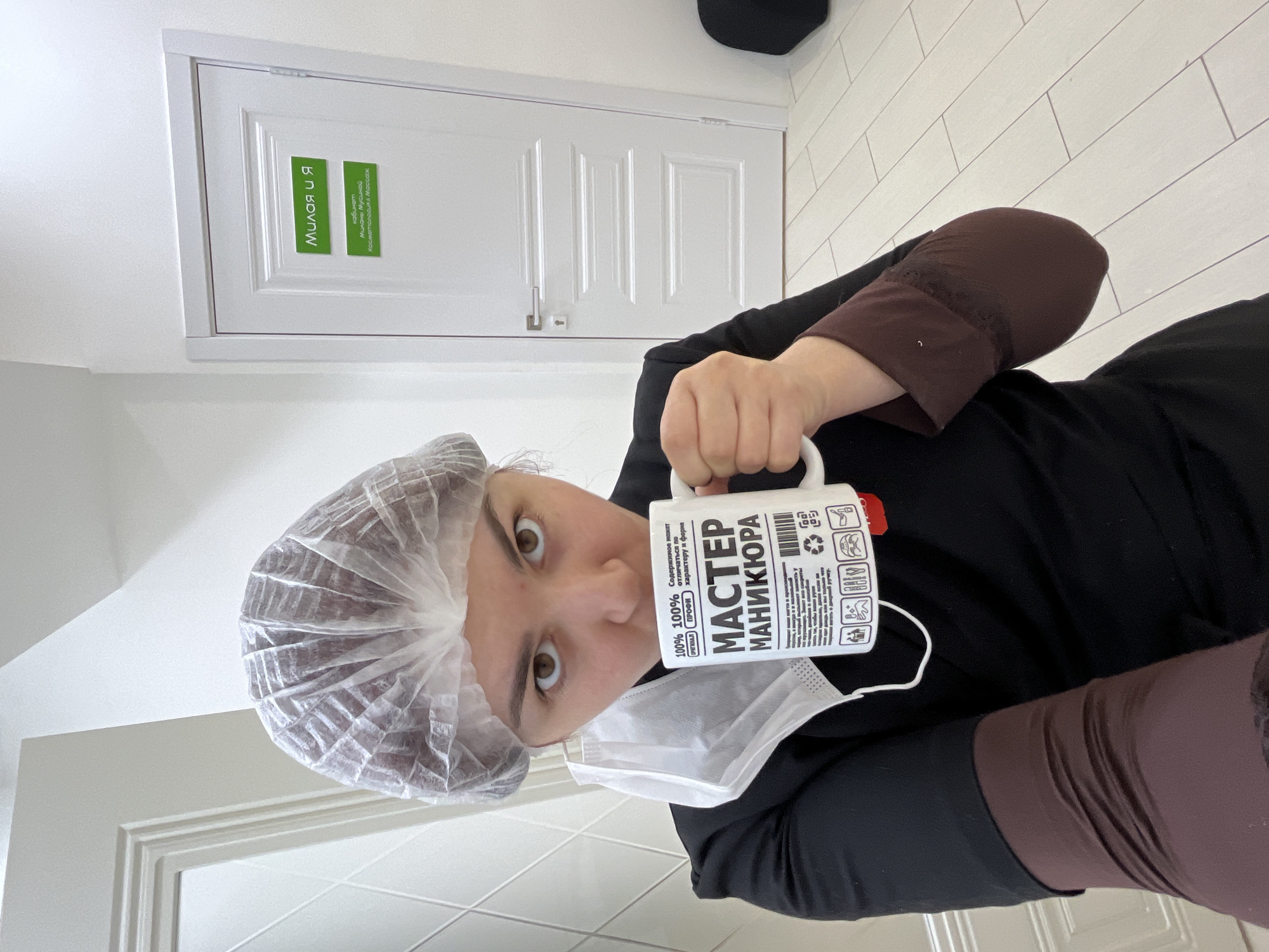 An individual is depicted indoors, within a brightly lit room featuring white walls and white paneled doors. The floor is covered with light-colored, possibly white or light grey, rectangular tiles arranged in a linear pattern.
The person is a female, looking directly at the camera. She wears a transparent, disposable hairnet. A white surgical face mask hangs from her left ear, resting against her black long-sleeved top. Underneath the black top, a brown long-sleeved garment is visible. She holds a white ceramic mug with a red base in her right hand, bringing it towards her mouth. The mug bears Cyrillic text, including "МАСТЕР МАНИКЮРА" (MASTER OF MANICURE), and various small graphic icons.
Behind the individual, to the right, is a white paneled door with a silver handle and keyhole. Affixed to this door are two green rectangular signs. The upper sign reads "МИЛАЯ И Я" (MILAYA AND I). The lower sign reads "кабинет МИЛАНЫ МУСИНОЙ Косметология & Массаж" (cabinet MILANA MUSINA Cosmetology & Massage). These signs indicate the establishment is a cosmetology and massage cabinet belonging to Milana Musina, potentially part of a larger business named "Milaya and I." A partially visible, dark-colored object is situated on the floor to the far right. The presence of these signs, coupled with the user-provided context, identifies the location as a cosmetology and massage cabinet in the city of Neftekamsk, Russia.