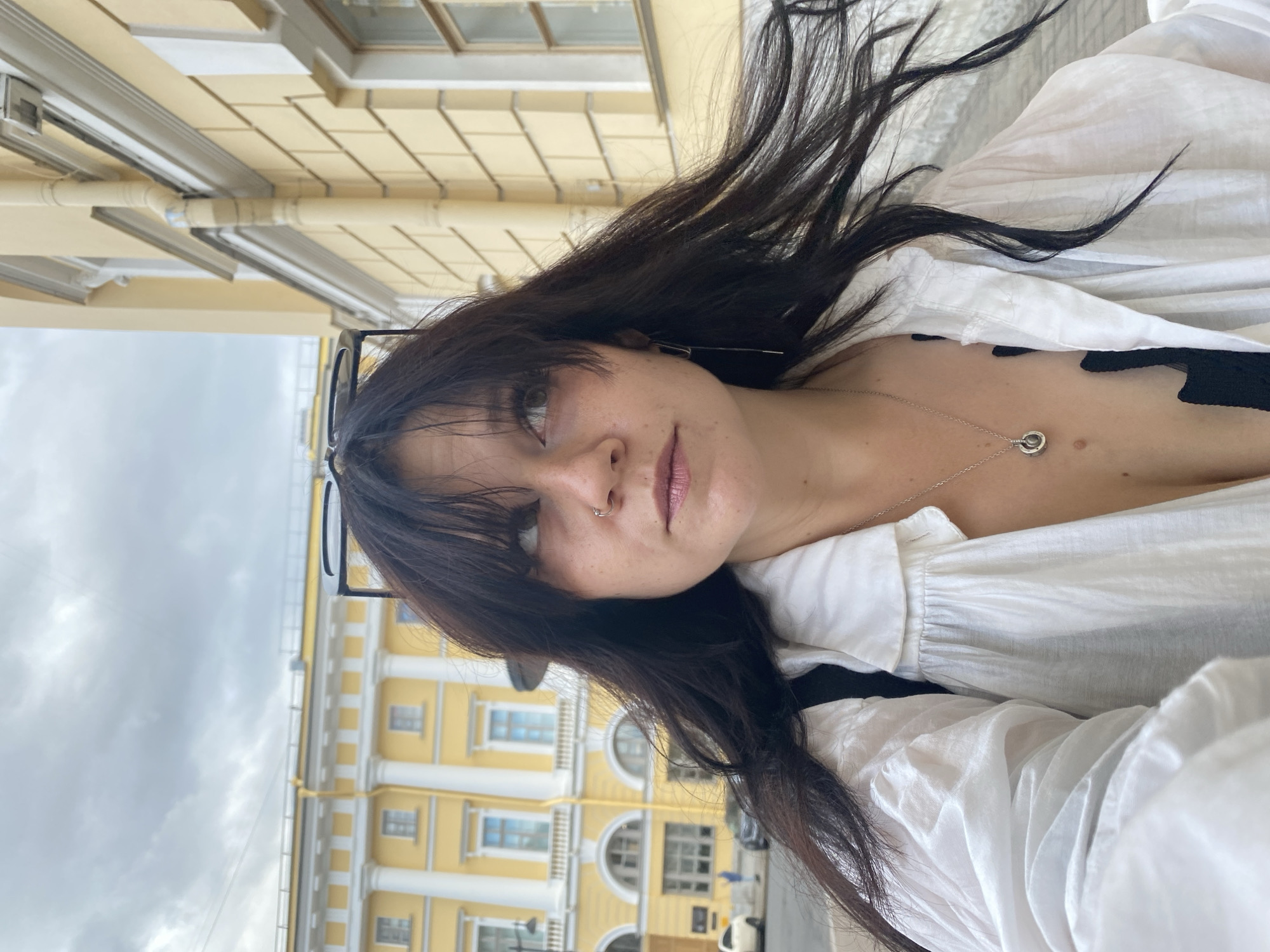 A medium shot captures a woman with long, dark, wind-blown hair, looking upwards and to her right. She is wearing a white open-collared shirt, a black top, and a silver necklace with a pendant. Her face is slightly turned away from the camera, revealing a nose ring and freckles. Behind her, a yellow building with white columns and arched windows suggests a location in Saint Petersburg, Russia, likely an architectural landmark with classical influences. The sky is overcast.