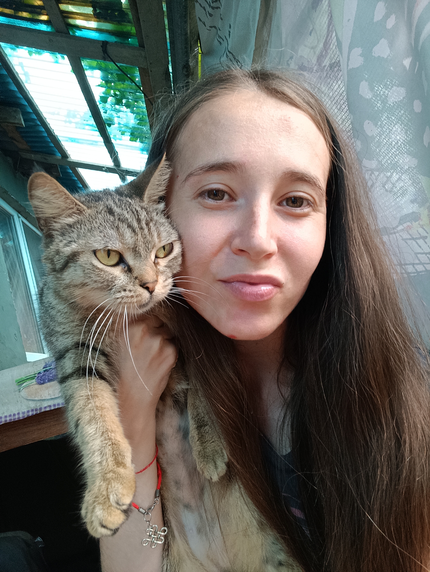 A young woman with long, dark brown hair is taking a selfie while holding a tabby cat. The woman is looking at the camera with a slight smile, and the cat is resting its head on her shoulder, looking to the side. They are in an outdoor setting, likely a veranda or covered patio, with a corrugated metal roof and some greenery visible in the background. The woman is wearing a black shirt and a red bracelet with a silver charm. The cat has green eyes and prominent whiskers. The overall scene suggests a casual, personal moment captured outdoors. The location is Vypasne, Ukraine.