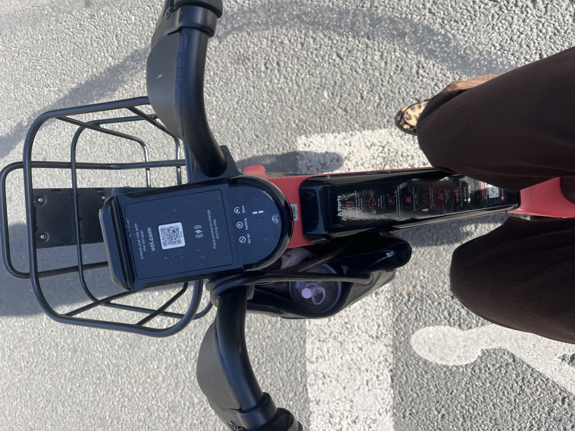 This image depicts the handlebars and front portion of a Voi electric scooter, likely in Paris, France, judging by the "À Paris" text on the scooter's display panel. The scooter has a black front basket. A black display panel with a QR code for the Voi app is mounted on the handlebars. The panel also has symbols for charging, speed controls (slow), NFC, no-go zones, and parking. The scooter is red. A person is near the scooter, wearing brown pants and a shoe with an animal print. The scooter is on an asphalt surface.
