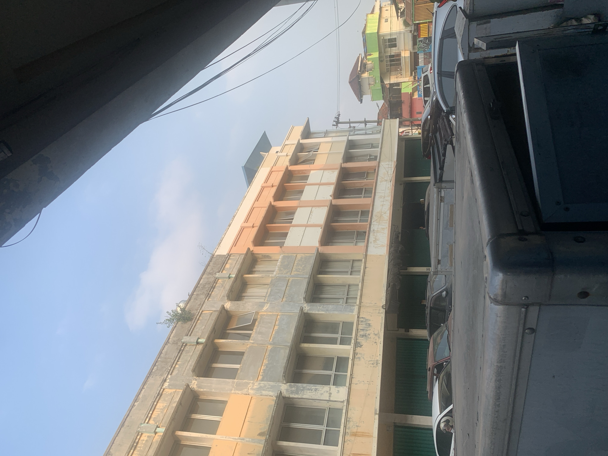 The image depicts a street scene in Palembang, Indonesia, focusing on a row of multi-story buildings with aging facades. The architecture suggests a commercial or residential area. The buildings are varied in color, with some sections painted in light orange and others in a weathered grey concrete. Numerous windows, many with closed curtains, line the upper floors. The ground floors appear to be shops or storefronts with green metal shutters. In the foreground, the side of a large, dark grey, metallic vehicle, possibly a truck or van, dominates the lower part of the frame, its textured surface and visible rivets indicating a utilitarian design. Behind this vehicle, several cars are parked or moving along the street, including a white SUV, a dark-colored SUV, and a maroon car. Beyond the initial row of buildings, other structures are visible, including brightly colored houses with pitched roofs, suggesting a densely populated urban environment. The sky is a pale blue with some wispy clouds, and the lighting indicates it is either early morning or late afternoon. Electrical wires are strung across the scene, and a lone tree is growing from the roof of one of the buildings.