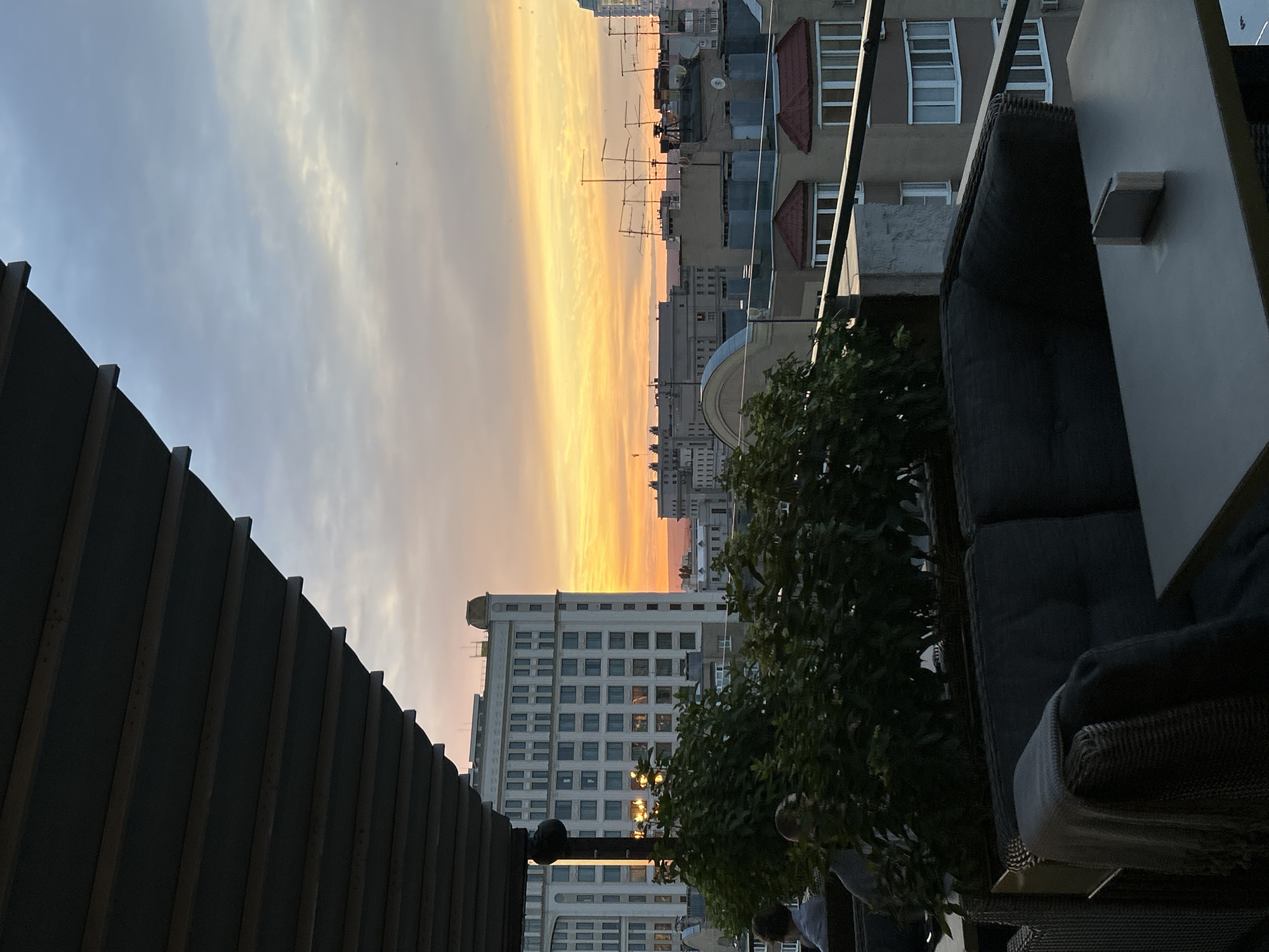 The image captures a sunset view from a rooftop or balcony in Novokuybyshevsk, Russia. In the foreground, an outdoor seating area is visible, featuring a dark gray sofa with a blanket draped over it and a table with a small rectangular object on it. Beyond the seating area, lush green plants form a dense hedge. The background is dominated by a cityscape with various buildings. A prominent, light-colored multi-story building with numerous windows stands out. Other buildings with tiled roofs and antennas are also present. The sky is a spectacle of sunset colors, with hues of orange, yellow, and pink blending into a pale blue. The lighting suggests it is late evening. Two individuals are partially visible sitting on a bench behind the plants, appearing to be observing the sunset.