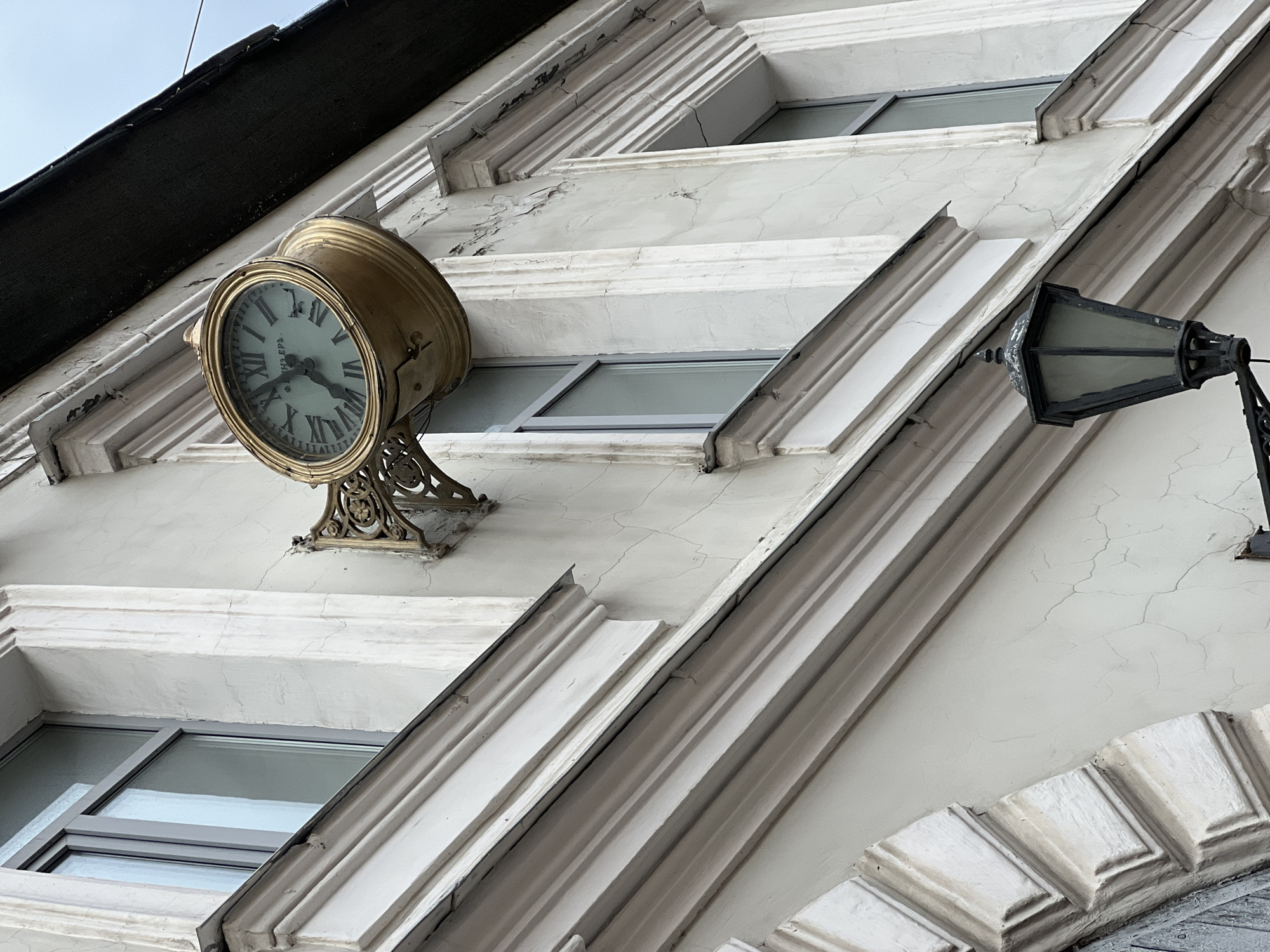 A low-angle shot showcases a building facade in Saint Petersburg, Russia. The architectural details include windows framed by ornate, white cornices. A gold-colored clock, displaying Roman numerals, is affixed to the wall. A black, vintage-style lamppost is mounted on the right side of the building. The building's exterior exhibits a weathered appearance with cracks in the paint.