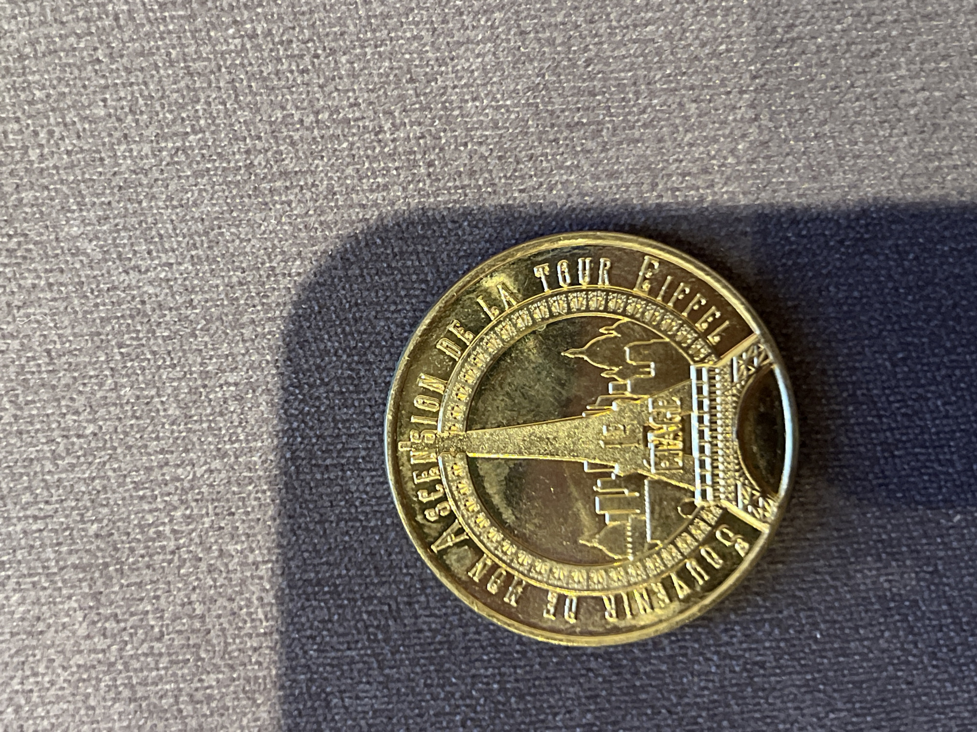 The image is a close-up, overhead shot of a gold-colored commemorative coin resting on a textured gray surface. The coin features an embossed design depicting the Eiffel Tower and surrounding buildings, along with French text that reads "SOUVENIR DE NON ASCENSION DE LA TOUR EIFFEL" and "1 ER ETAGE". The coin is circular with an ornate border. The gray surface appears to be fabric, possibly upholstery or a rug, with visible texture and a shadow cast across the bottom left quadrant of the image. There are no people or actions depicted in the image. The coin is the sole subject. There is no information in the image to suggest a location in Montfermeil, France.