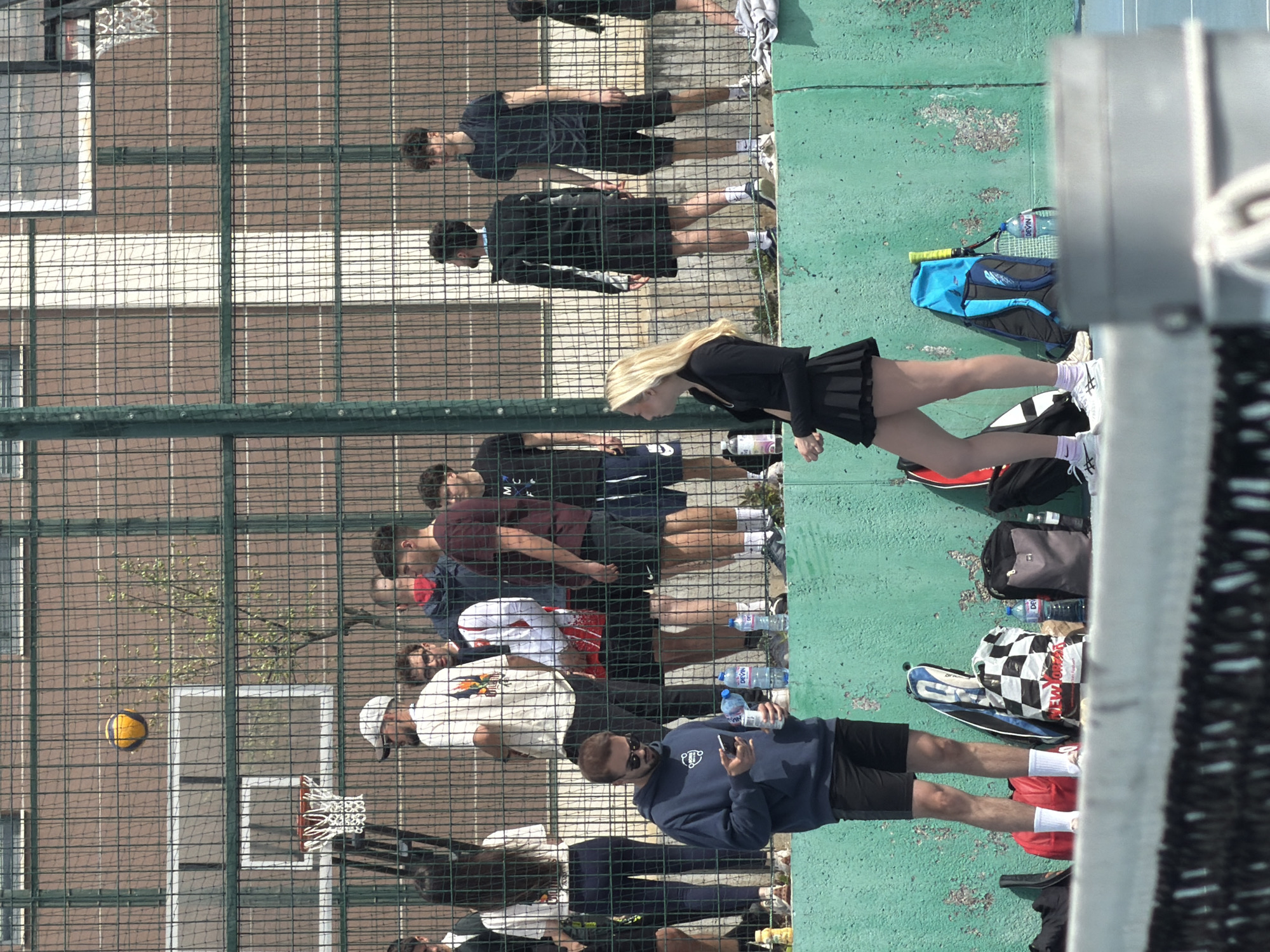 A medium-angle, outdoor shot shows multiple people standing next to a chain-link fence, seemingly at a tennis court located in Blagoevgrad, Bulgaria. Several of the people are dressed in athletic attire, while others wear casual clothing. A basketball hoop is visible through the fence. A basketball appears to be suspended in the air near the hoop. Bags and water bottles are placed on the green surface. One person is looking at their cell phone. The setting appears to be a sports complex.