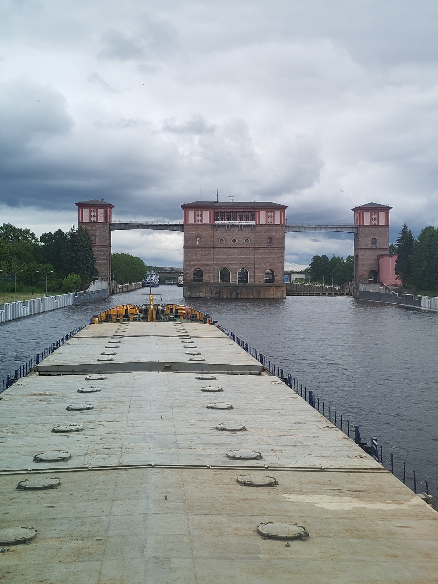 A wide-angle, eye-level shot shows a large barge moving towards the Rybinsk Lock in Rybinsk, Russia, under an overcast sky. The barge deck, light grey and marked with numerous circular hatches, takes up the foreground. Behind the barge, the water stretches towards the lock.
The Rybinsk Lock is constructed of red brick and is composed of three main structures. Two towers flank the waterway and are connected by a bridge-like structure at their upper levels. The central structure houses the lock machinery and control mechanisms. The buildings have red roofs and light red decorations.
Green foliage and trees line the banks of the canal leading up to the lock. A row of white concrete posts marks the canal edge on the left side. A boat can be seen past the lock.