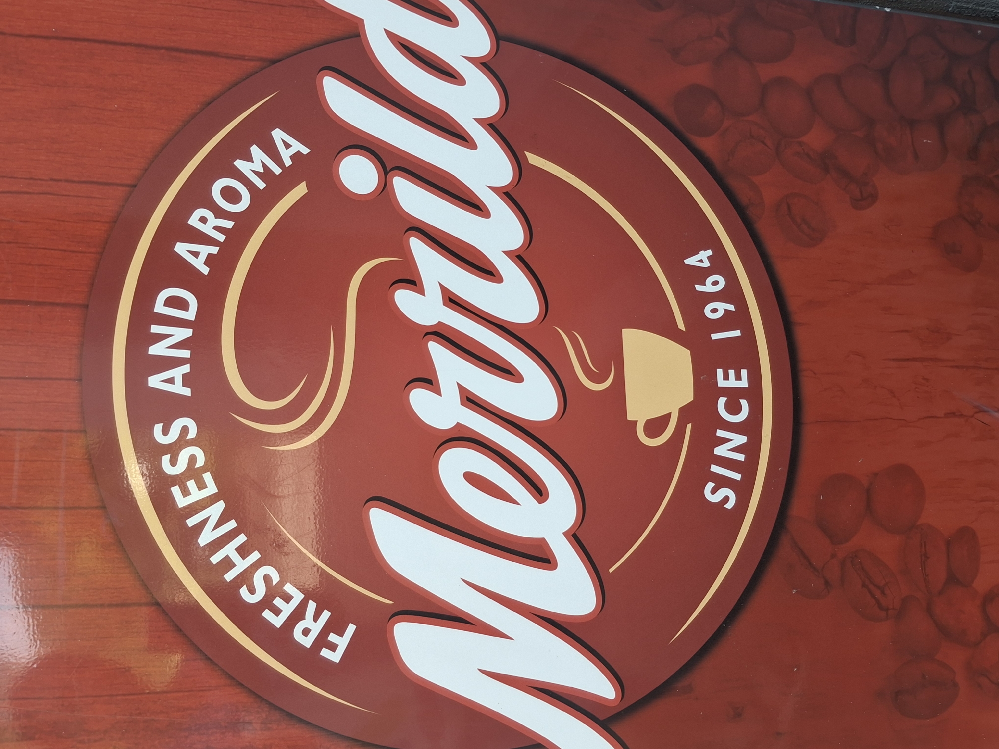 This is a close-up, eye-level photograph of a sign for "Merrild" coffee. The sign is predominantly red with a circular emblem in the center, which is also red and bordered by a thin yellow line. Within the emblem, white text reads "FRESHNESS AND AROMA" arched across the top. Below this, in large white script with a red outline, is the brand name "Merrild." Beneath "Merrild" is a stylized yellow image of a coffee cup with steam rising from it, and below that, white text reads "SINCE 1964." The background of the sign appears to be a wooden texture. The lower portion of the image shows a scattering of coffee beans, also in a reddish hue. There are no people or animals visible in the image. The location in Lēdmane, Latvia, is not discernible from the image itself.