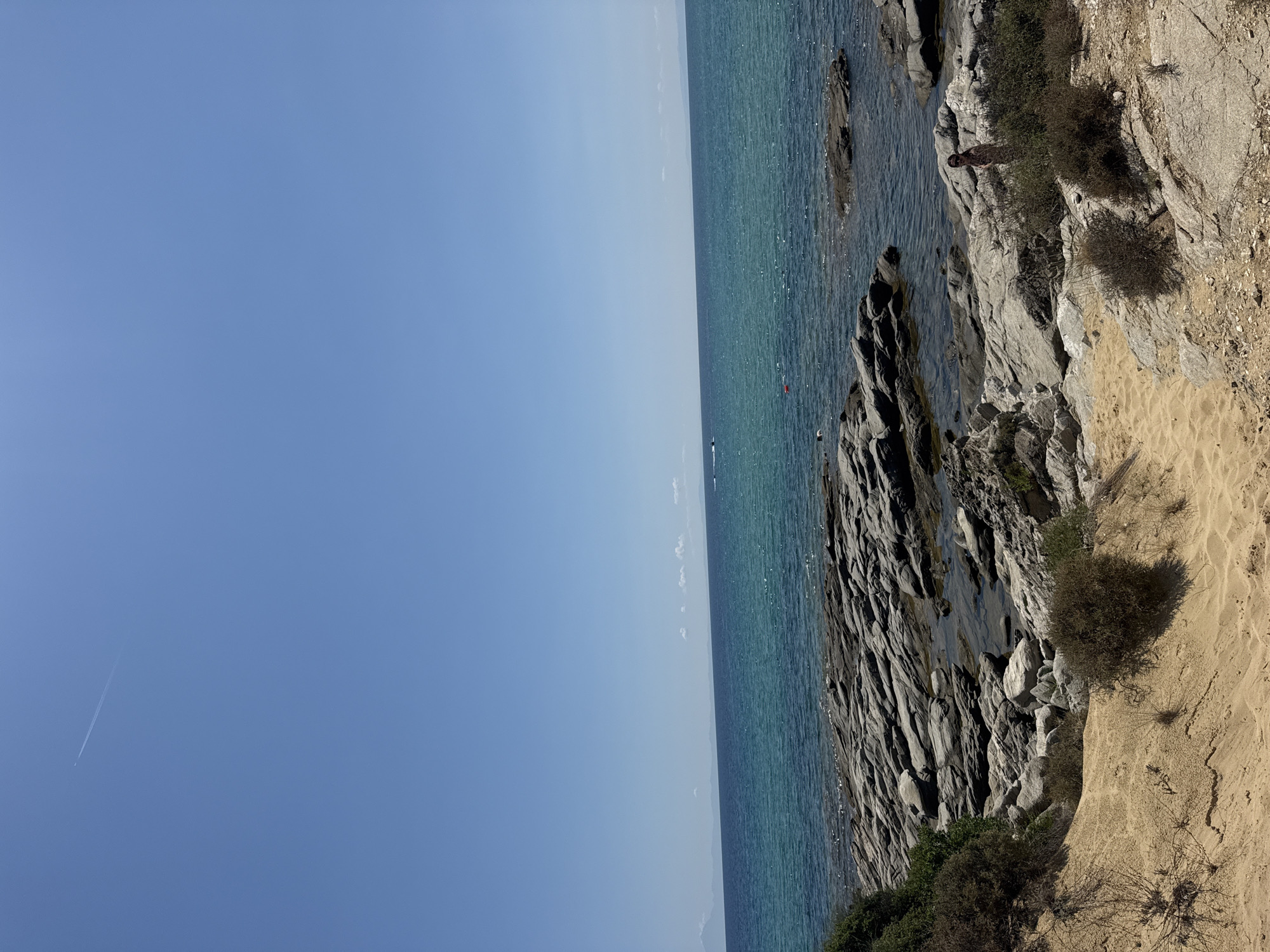 A wide-angle shot captures a serene coastal scene in Nea Peramos, Greece. The sky is a clear, expansive blue, with a faint contrail from an airplane visible in the upper left corner. Below, the turquoise waters of the Aegean Sea gently lap against a rocky shoreline. The foreground features a sandy path leading down towards the water, bordered by dry, scrubby bushes. Large, grey rocks are scattered along the beach and in the shallow water, some smooth and others jagged. In the distance, the coastline is visible as a faint, hazy line against the horizon. A small red buoy floats in the water. A lone individual, partially obscured by vegetation on the right side of the frame, stands amidst the rocks, appearing to be observing the sea.