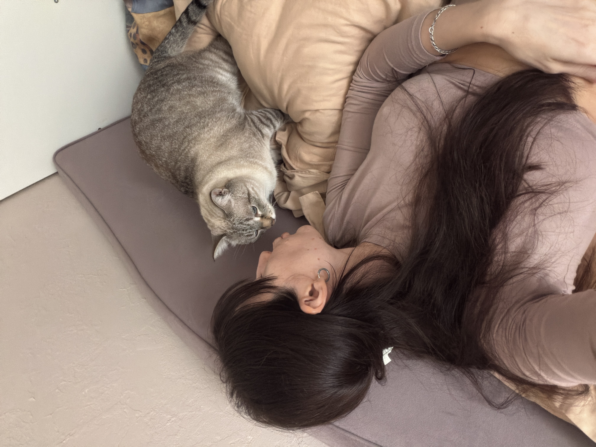 A woman with long, dark hair is lying on a grey mattress, possibly a bed. She is wearing a light pink or beige long-sleeved shirt and a silver bracelet on her left wrist. Her head is turned to the left. An earring is visible on her left ear.
A grey tabby cat is positioned near the woman's head, likely on the same mattress. The cat is facing the woman and appears to be interacting with her.
A beige pillow is partially visible between the woman and the cat. The location is presumed to be Perm, Russia. The environment consists of the interior space, which includes the mattress, wall, and flooring.