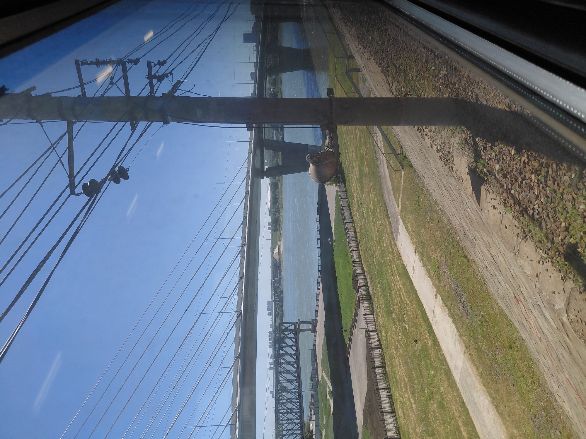 This is a photograph taken from a moving vehicle, likely a train, looking out a window. The view is of a wide river in Novosibirsk, Russia, with two bridges spanning it. The foreground is dominated by a utility pole with numerous wires attached. To the left of the pole, there is a grassy embankment and a concrete retaining wall. Beyond the embankment, a river is visible with a large, cable-stayed bridge crossing it in the background. In the middle ground, a smaller, older truss bridge is also visible. The sky is clear and blue. The image is slightly distorted due to the window of the vehicle.