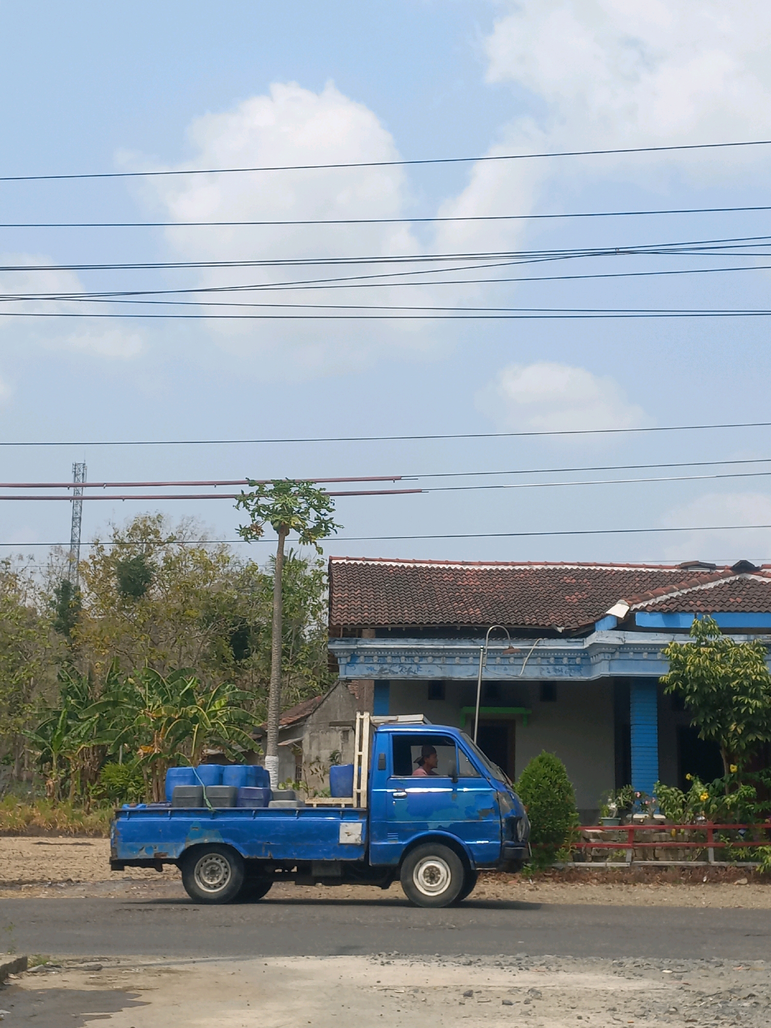 In Baturetno, Indonesia, a blue pickup truck drives on a paved road. The truck bed is loaded with several blue and gray barrels. A person is visible driving the truck. In the background, a single-story house with a tiled roof is present, along with trees and vegetation. Power lines are strung across the sky, which is partially cloudy. A cell tower is visible in the distance.