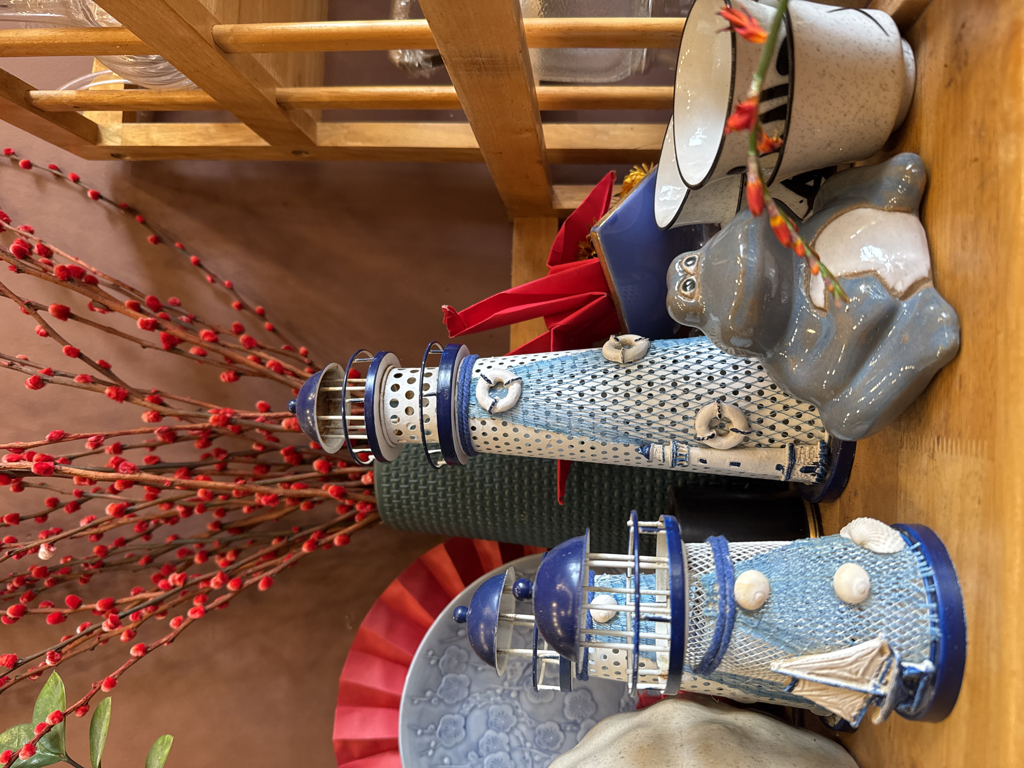 A still-life arrangement on a wooden shelf features nautical-themed decorations. Two lighthouse miniatures, painted in blue and white with shell and net embellishments, are centrally placed. Nearby is a ceramic figurine of an animal, possibly a hippopotamus, in shades of gray and blue. Also visible are a few ceramic mugs or cups in white with black accents. A fan partially obscured by the decorations and a vase filled with red berry branches are situated on the left side of the arrangement. The location is likely in Nha Trang Ward, Vietnam.