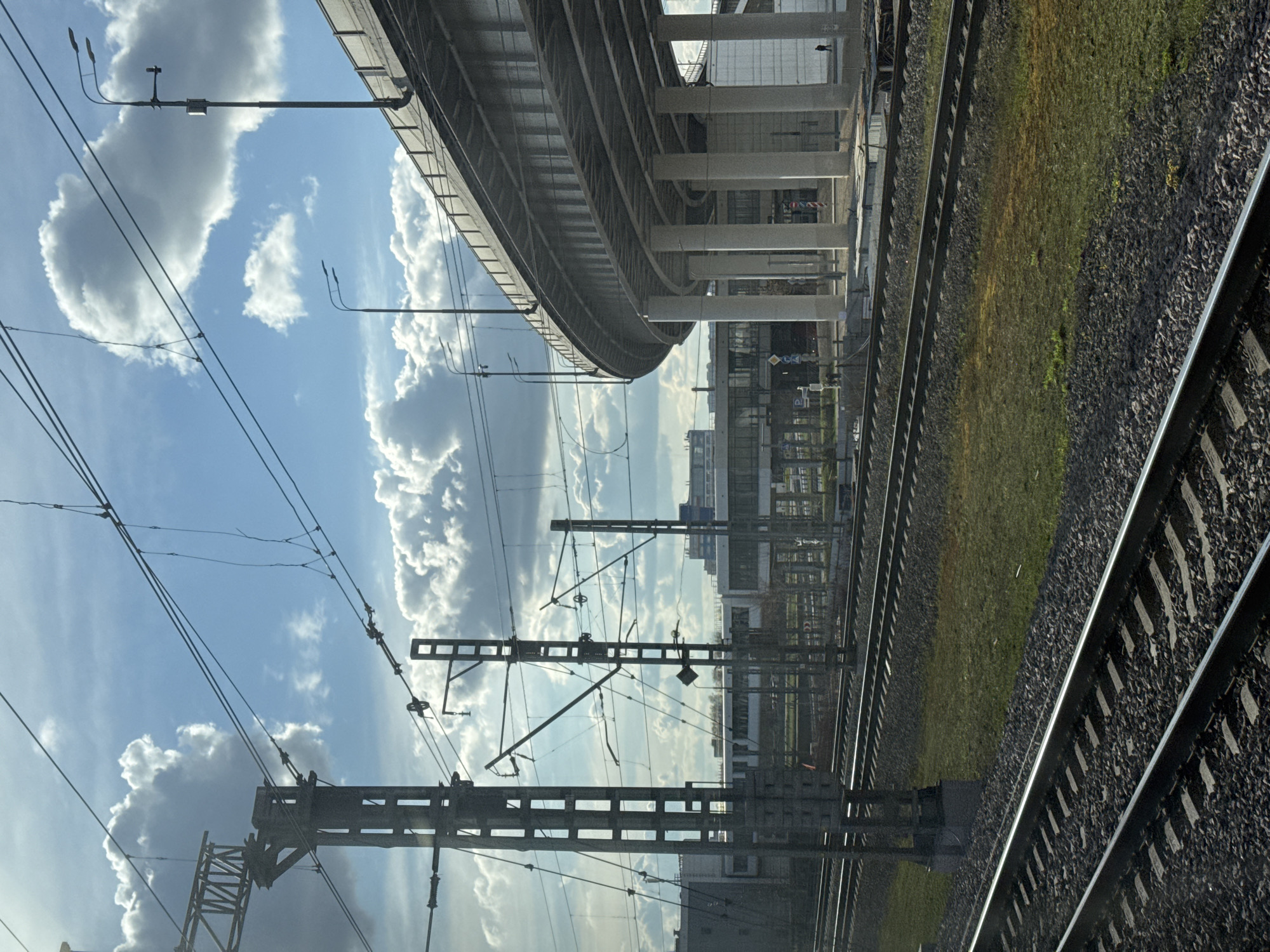 The image depicts a view from a train window in Khimki, Russia. Railway tracks run parallel to each other in the foreground. Adjacent to the tracks is a patch of green vegetation. Overhead electrical wires and support structures for the railway's power system are prominent against a blue sky with scattered cumulus clouds. An elevated road bridge curves overhead, supported by white concrete pillars. Beyond the tracks and bridge, indistinct buildings are visible in the background. The sky dominates the scene.