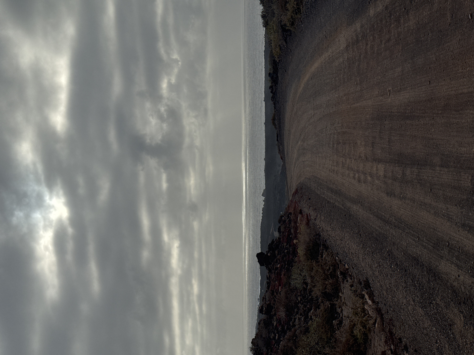 The image captures a view from the rugged coastline of Tinajo, Lanzarote, Spain. On the left is a sky filled with textured, gray clouds, partially obscuring the sun, creating luminous patches. A strip of the Atlantic Ocean separates the sky from the land. The ocean surface has a band of sunlight reflecting off it. The right side of the image is dominated by a brown dirt path or road curving along a rocky terrain. The ground is composed of dark, volcanic rocks and sparse vegetation.