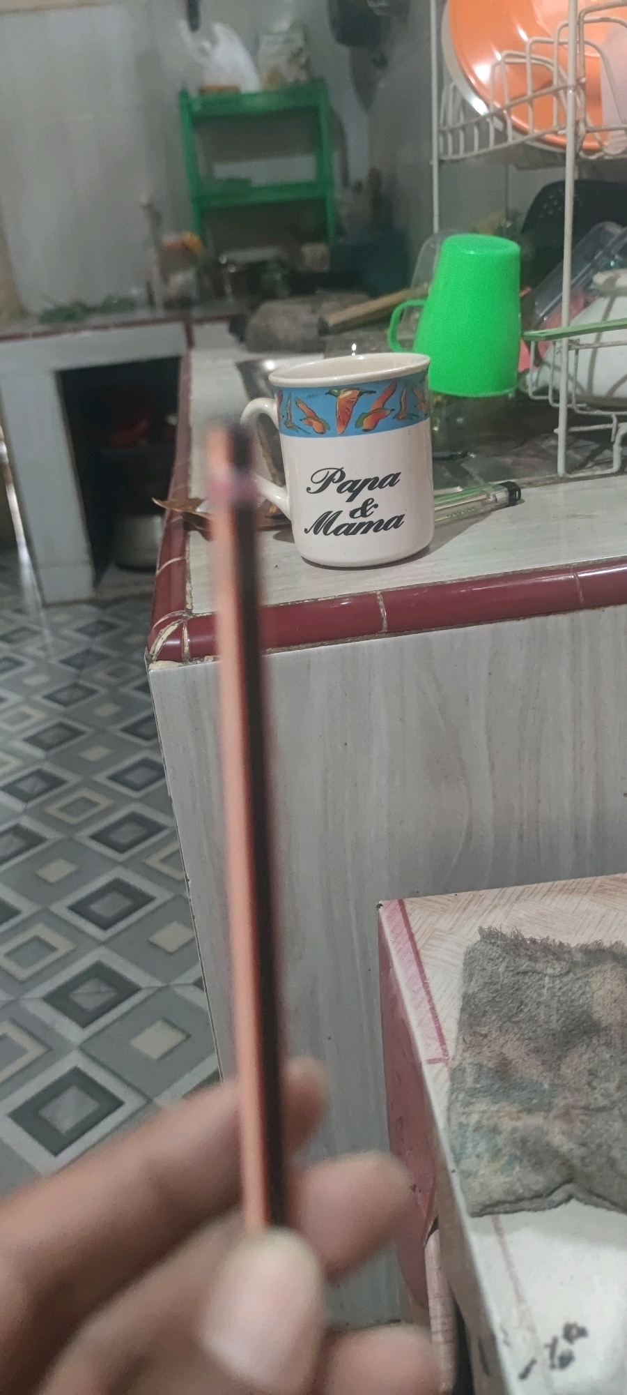 The image shows a close-up view of a hand holding a pencil, with a mug on a countertop in the background. The mug is white with a blue and orange bird pattern around the rim and the words "Papa & Mama" written in black script below the pattern. Next to the mug, a green plastic cup is visible, as well as part of a white dish rack with orange plates. The countertop is tiled with a red and white pattern. The background shows a kitchen area with shelves and various objects, including what appears to be a dish drying rack. The floor has a diamond-patterned tile. The lighting is dim, suggesting an indoor setting. The overall scene appears to be in a domestic environment. There is no information in the image that can be used to determine the location within Gambarsari, Indonesia.