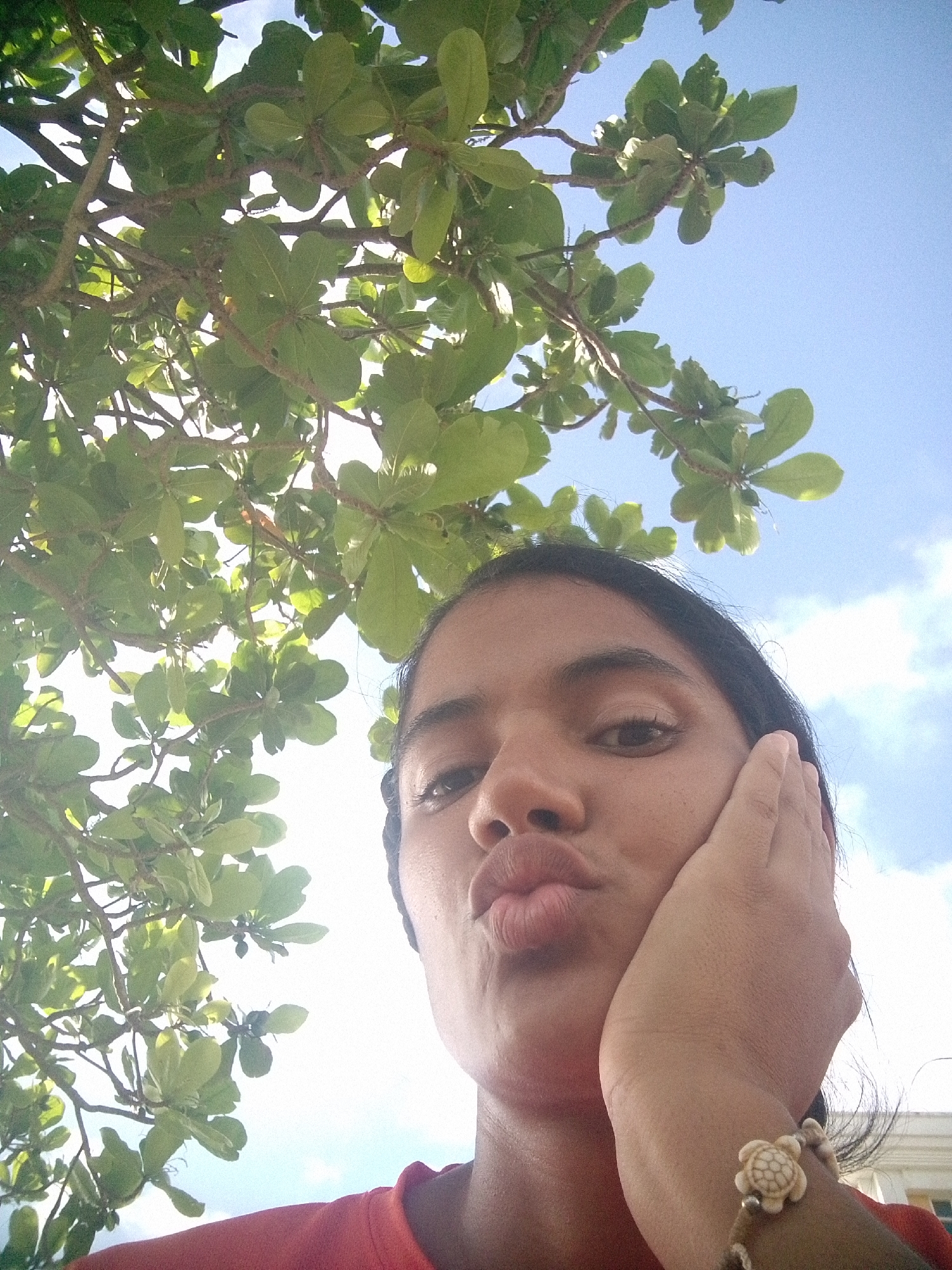 A young woman, likely of South Asian descent, poses for a selfie outdoors under a tree. She is looking at the camera with her lips pursed. Her right hand is resting on her cheek, and she is wearing a bracelet with a turtle charm. The background shows a blue sky with some clouds and the branches and green leaves of a tree. The image appears to have been taken in the city of Pedro González, Venezuela.