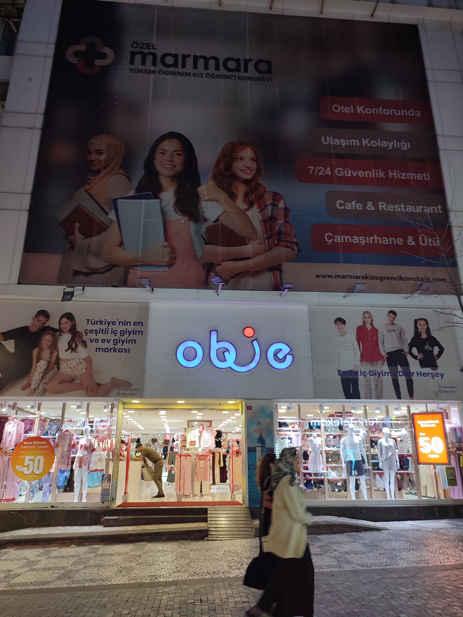 This is a nighttime exterior shot of a building with multiple advertisements and storefronts visible. Dominating the upper portion of the image is a large billboard for "Özel Marmara Yüksek Öğrenim Kız Öğrenci Konukevi" (Marmara Private Girls' Student Dormitory). The billboard features images of young women and text in Turkish, listing amenities such as "Otel Konforunda" (In hotel comfort), "Ulaşım Kolaylığı" (Easy transportation), "7/24 Güvenlik Hizmeti" (24/7 security service), "Cafe & Restaurant", and "Çamaşırhane & Ütü" (Laundry & Ironing).
Below the dormitory advertisement is a storefront displaying clothing. A prominent blue neon sign reads "obje". To the left of the sign, another advertisement features a family and text in Turkish stating "Türkiye'nin en çeşitli iç giyim ve ev giyim markası" (Turkey's most diverse underwear and loungewear brand).
The storefront itself has large glass windows showcasing mannequins dressed in sleepwear and loungewear. Various clothing items are displayed on shelves and racks. A prominent orange sign displays "%50" indicating a discount. A man in a green shirt is seen inside the store, interacting with a counter.
To the right of the "obje" sign, another advertisement features a family and text that appears to be related to children's clothing. Further to the right, there are more displays of clothing and mannequins.
The ground in front of the building is paved with cobblestones. In the foreground, two women are walking past the storefront. The overall lighting suggests it is nighttime, with the illuminated advertisements and storefronts creating a bright contrast against the dark surroundings. The architectural style appears modern. The building is likely located in the Kadıköy district of Istanbul, Turkey, given the Turkish text and common advertising styles for businesses in that area.