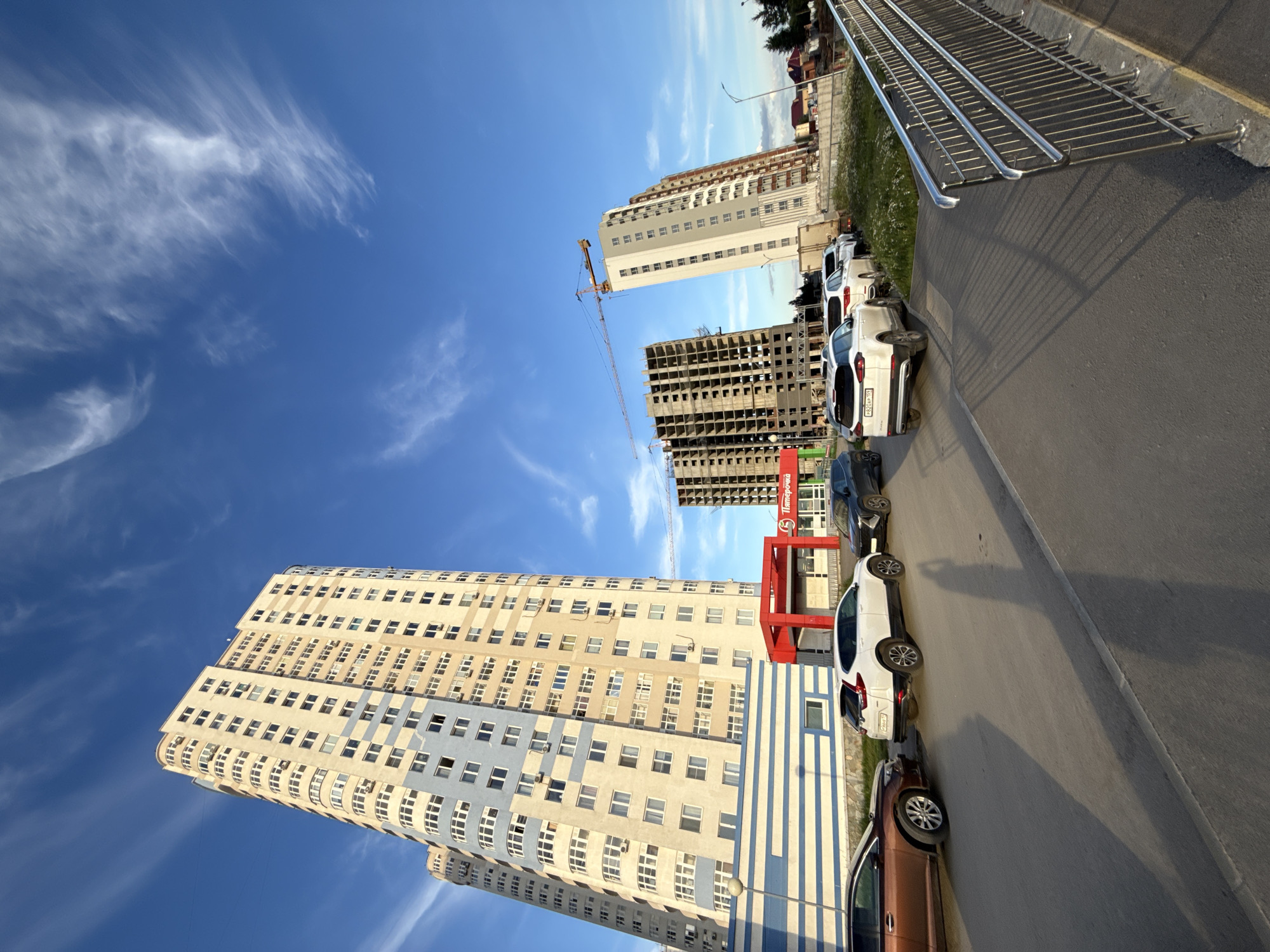 This media file captures an outdoor scene with a clear blue sky and wispy white clouds, indicating fair weather. The time of day appears to be late afternoon or early evening, as evidenced by the long shadows cast by the objects in the frame. The setting is an urban environment, likely in Perm, Russia, characterized by modern architecture and ongoing construction.
The primary subjects are several multi-story buildings, some completed and inhabited, others in various stages of construction. One prominent completed building is tall and slender, with a facade of beige and light blue horizontal stripes and numerous windows. Adjacent to it, a building under construction reveals its skeletal concrete structure. A construction crane is visible near this unfinished building.
In the foreground, a paved road runs alongside a grassy area and a metal railing, possibly part of an elevated walkway or bridge. Several cars are parked along the side of the road. The cars are white, black, and brown, and their license plates are visible, though not legible in detail. One white car has the license plate "B824AP 11".
A red architectural element, possibly an entrance or part of a storefront, is situated between the completed and under-construction buildings. Visible text on a green sign reads "Пятерочка" which translates to "Pyaterochka", a Russian supermarket chain. The overall impression is one of urban development and daily life in a Russian city. There are no people visible in the image.