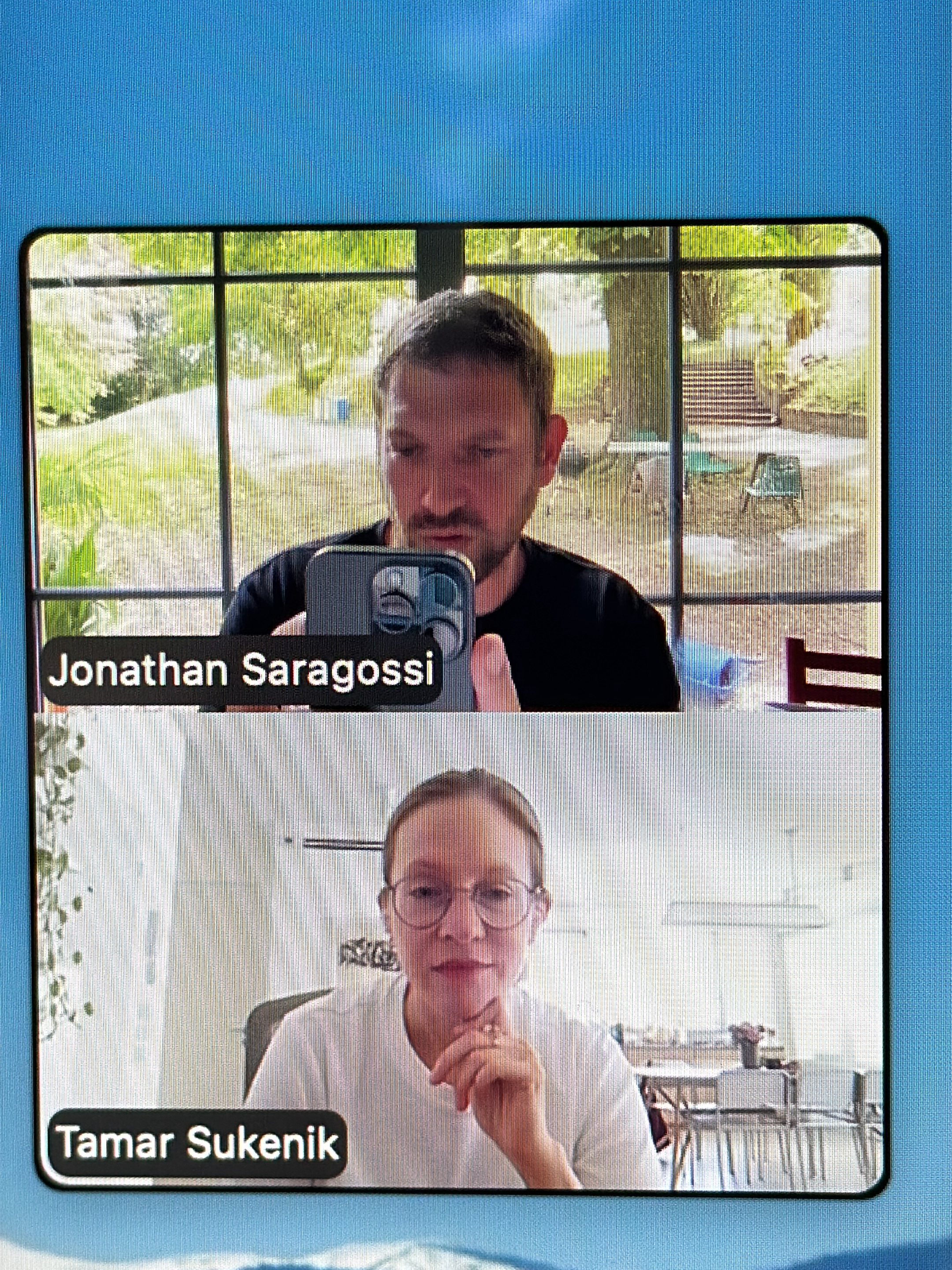 This media file is a screenshot of a video conference call, showing two participants in separate frames. The top frame features a man holding a smartphone, appearing to take a selfie or record a video. He is wearing a black t-shirt and has short brown hair and a beard. Behind him, a large window reveals a lush green outdoor setting with trees and what appears to be an outdoor seating area. The bottom frame displays a woman wearing glasses and a light-colored t-shirt. She has her hand near her chin, and her expression is neutral. The background behind her is an indoor setting, with tables and chairs visible. The overall lighting suggests it is daytime.
Visible text in the image includes "Jonathan Saragossi" in the bottom left corner of the top frame and "Tamar Sukenik" in the bottom left corner of the bottom frame. These likely represent the names of the participants in the video call. The blue background surrounding the video frames is typical of a digital interface.