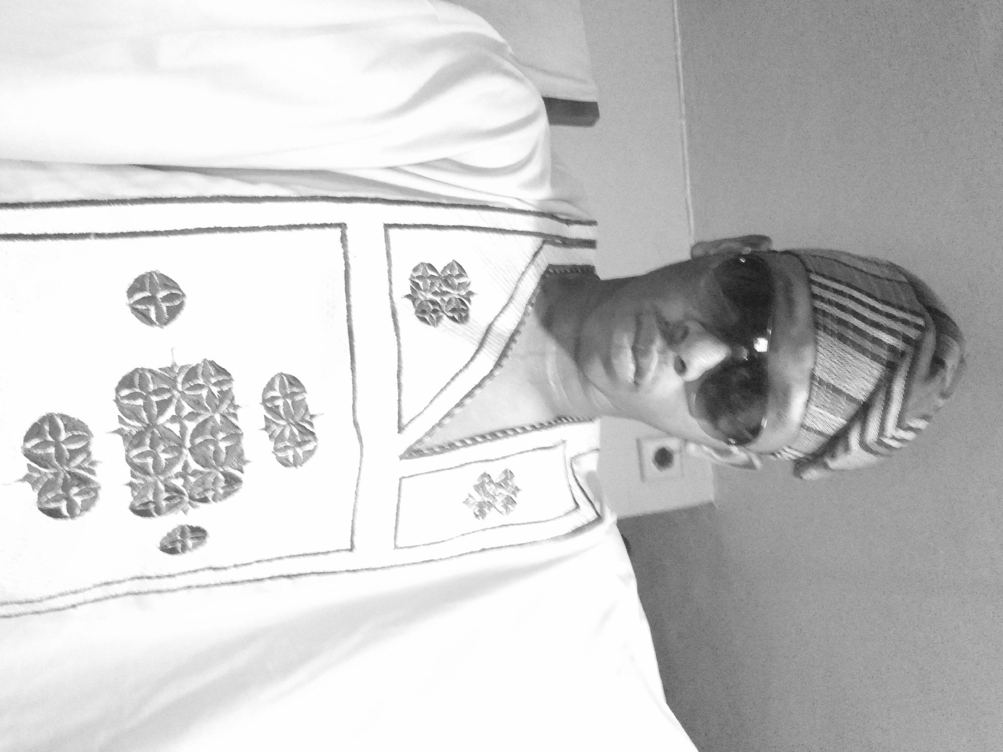 In Lagos, Nigeria, a man is captured in a medium shot, dressed in traditional attire. He wears a headwrap adorned with striped patterns and dark sunglasses. His garment, likely an Agbada, is white with intricate, dark embroidered detailing on the chest area, forming geometric patterns. He is looking directly at the camera, with a neutral expression. The background appears to be an indoor setting, with plain walls visible. The lighting is somewhat harsh, creating strong contrasts, and the image is in black and white, suggesting it may have been taken with a phone camera.