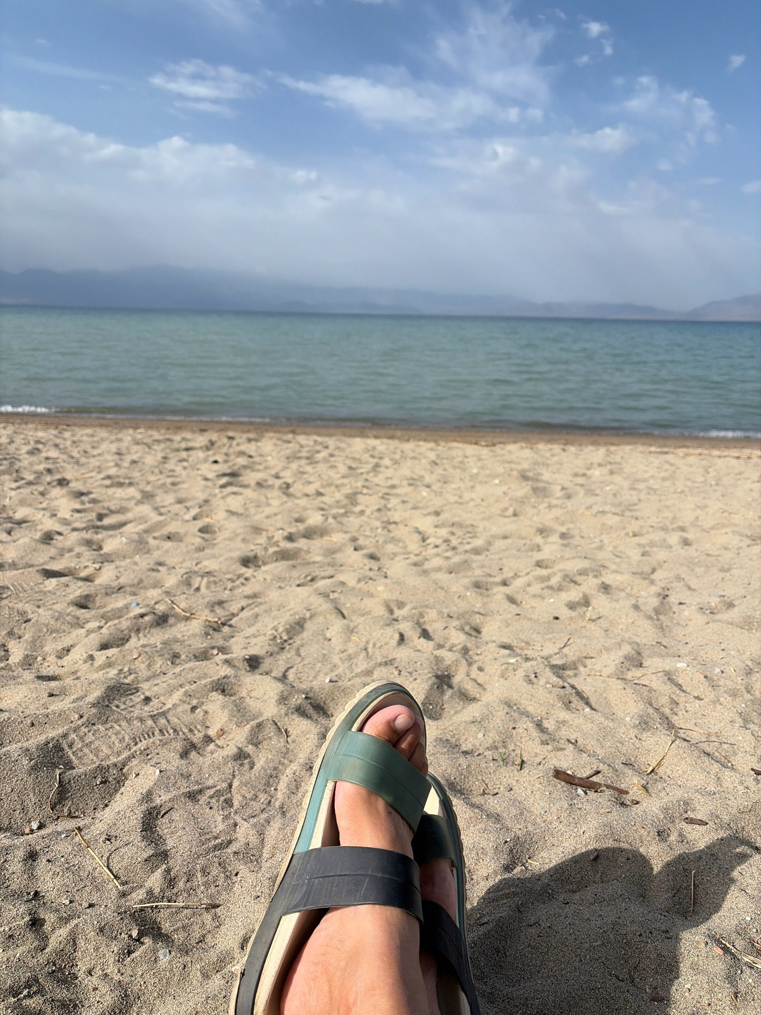 The image shows a first-person perspective of someone's feet wearing sandals, resting on a sandy beach. The background features a body of water, possibly a lake or ocean, with hazy mountains in the distance under a partly cloudy sky. The sand is light brown and appears somewhat dry with a few small pieces of debris. The water is a greenish-blue color. The scene suggests a relaxed, outdoor setting.
