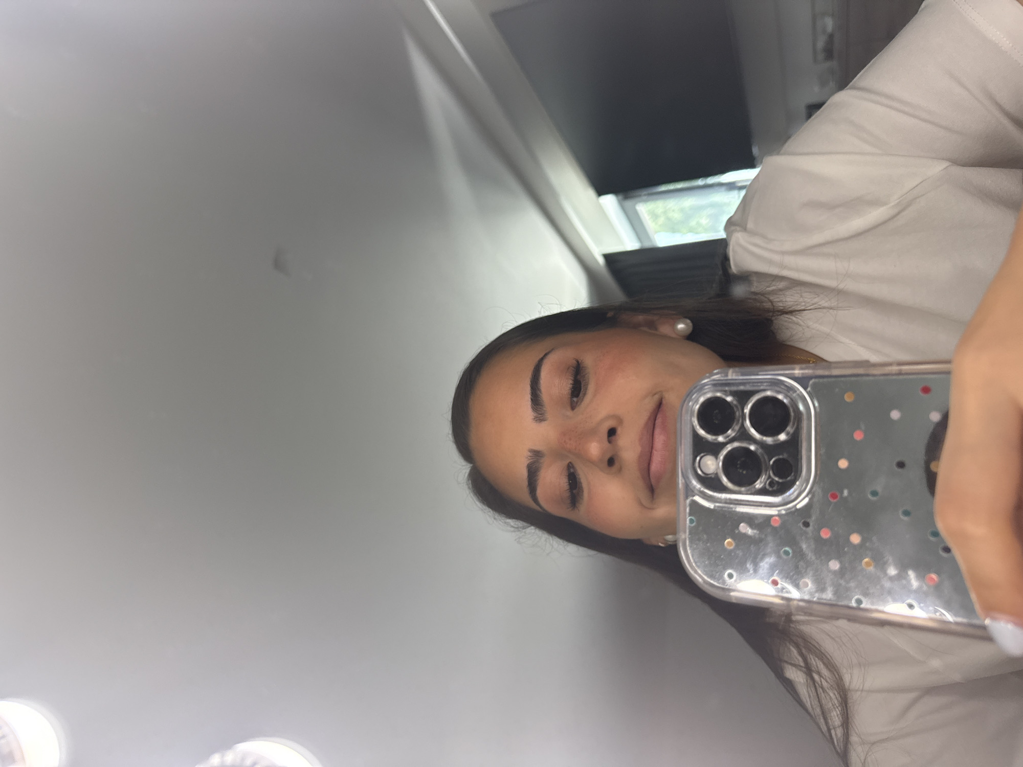 A person is taking a selfie indoors, with a gray wall and a window visible in the background. The person is holding a cell phone with a clear case decorated with colored dots. The individual has dark hair, fair skin, and is wearing a white top. The location cannot be confirmed as San Bernardino Tlaxcalancingo, Mexico.
