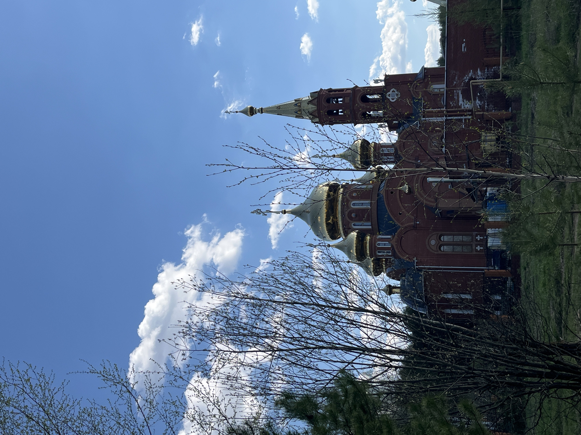 The image features the Holy Great Martyr George Victory-Bearer Cathedral in Chaikovsky, Russia, viewed from a low angle. The cathedral's red brick facade and golden domes are partially obscured by tree branches in the foreground. The sky is blue with scattered white clouds.