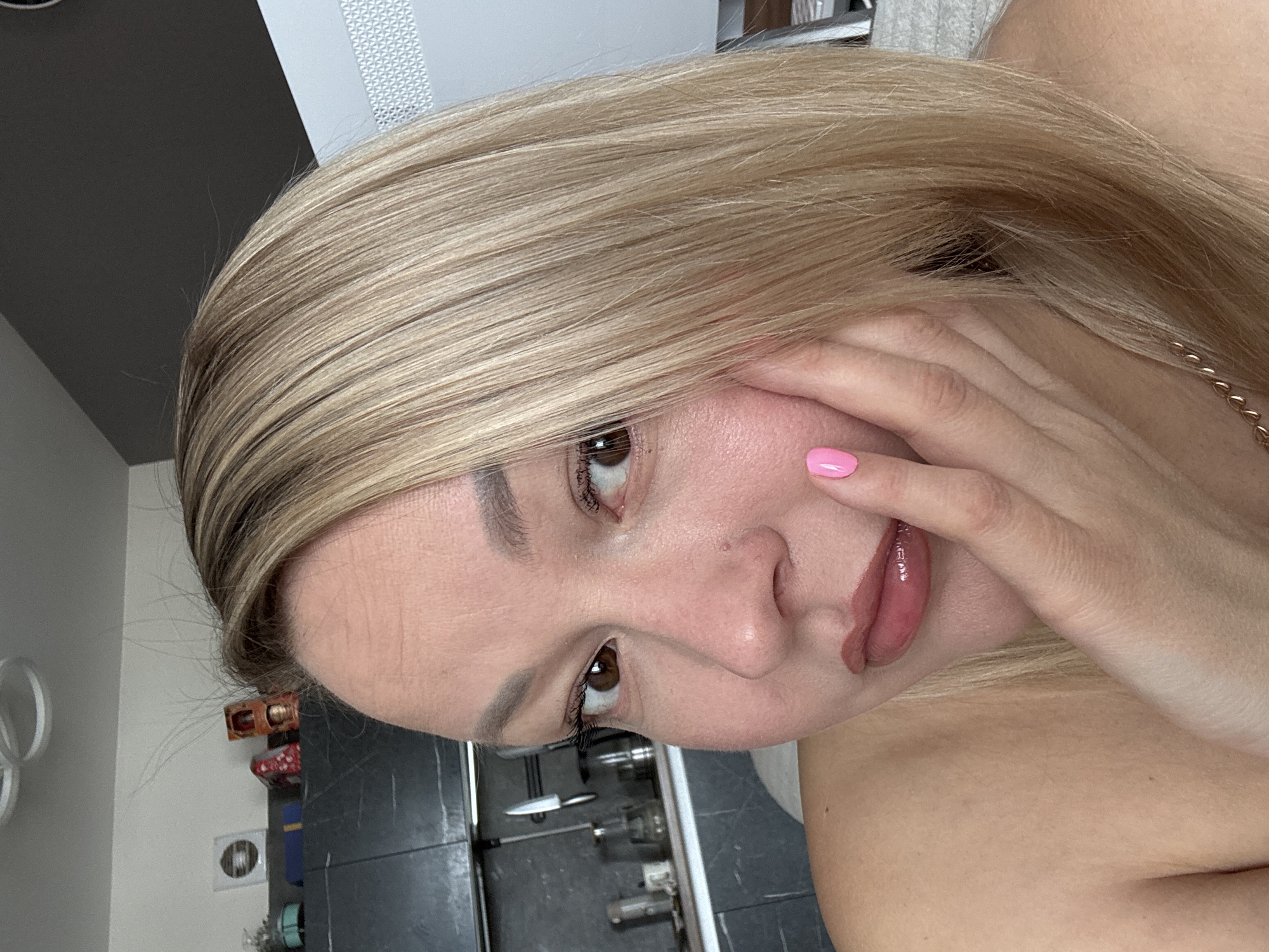 A close-up, slightly angled portrait of a light-skinned woman with blonde hair, brown eyes, and pink nail polish on one visible fingernail. The woman is resting her hand on her face. A gold chain necklace is partially visible on her neck. The background includes a kitchen countertop with kitchen items, potentially a dishwasher, and a wall with decorations. There is no discernible information to ascertain this image was taken in Izhevsk, Russia.