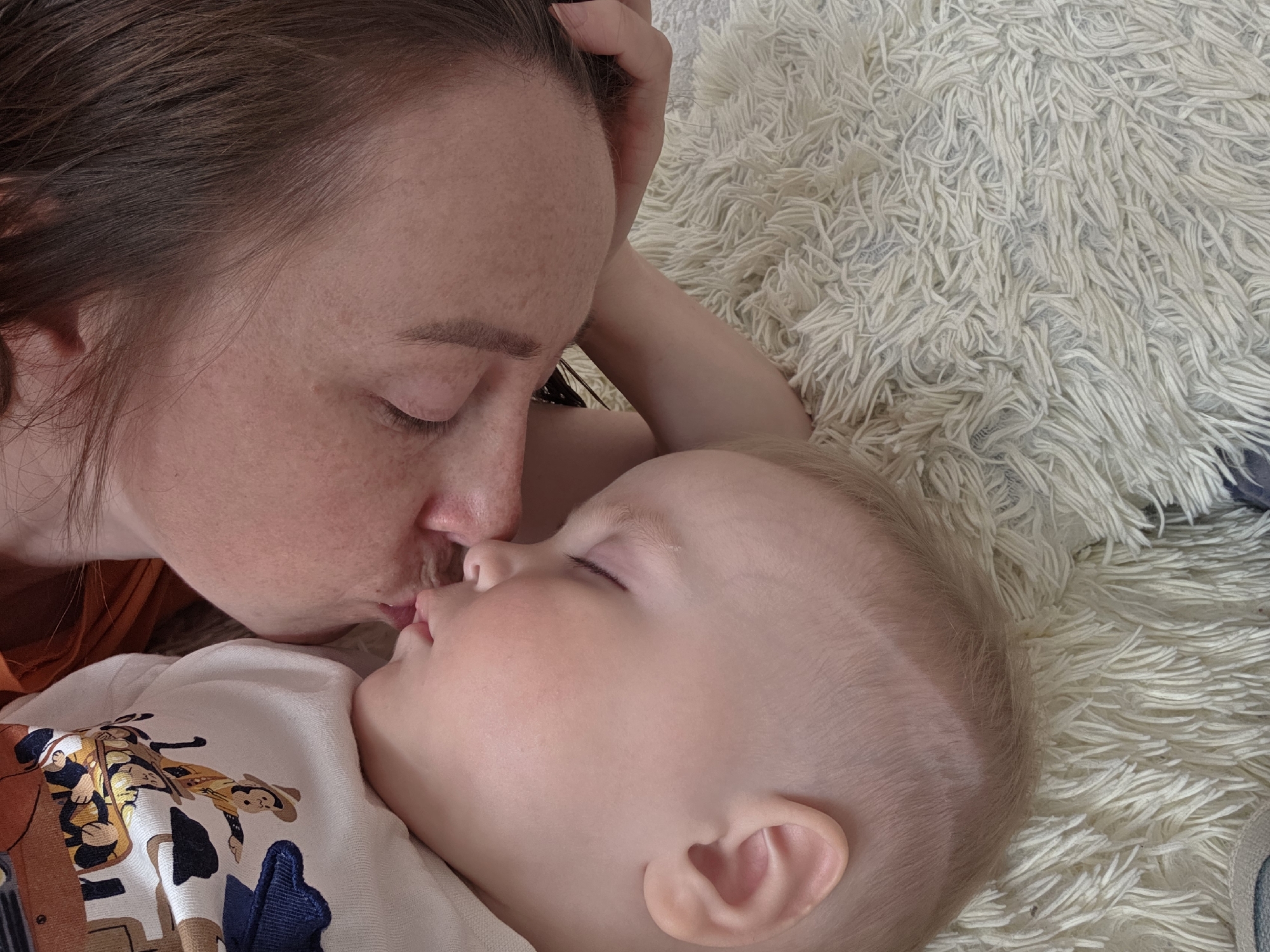 A woman and a baby are lying on a light-colored, shaggy rug. The woman is on her side, facing the baby, and is kissing the baby on the lips. The baby is on its back, eyes closed, wearing a white shirt with a cartoon graphic. The woman has brown hair and closed eyes. The background consists only of the shag rug. The location is Chaikovsky, Russia.