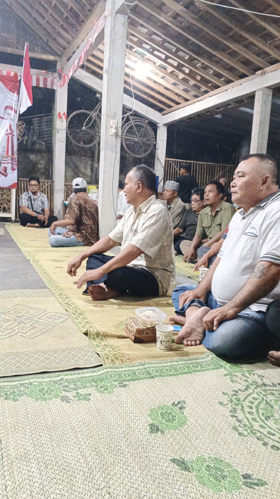 In Ambarketawang, Indonesia, a group of men are seated on woven mats under a covered, open-air structure with a wooden roof. The setting appears to be a gathering or event. Two bicycles are suspended from the ceiling. Several men are in the foreground, facing to the left. One man in a light-colored checkered shirt is speaking. Another man in a white polo shirt with a stripe on the collar is in the right foreground. Behind them, other men are also seated, some wearing traditional caps. Red and white decorative banners are visible on the left. The ground is covered with various woven mats, one with a prominent green floral pattern. A small box containing food and a cup are on the mat in the foreground. The overall lighting is dim, suggesting it may be evening or indoors with artificial light.