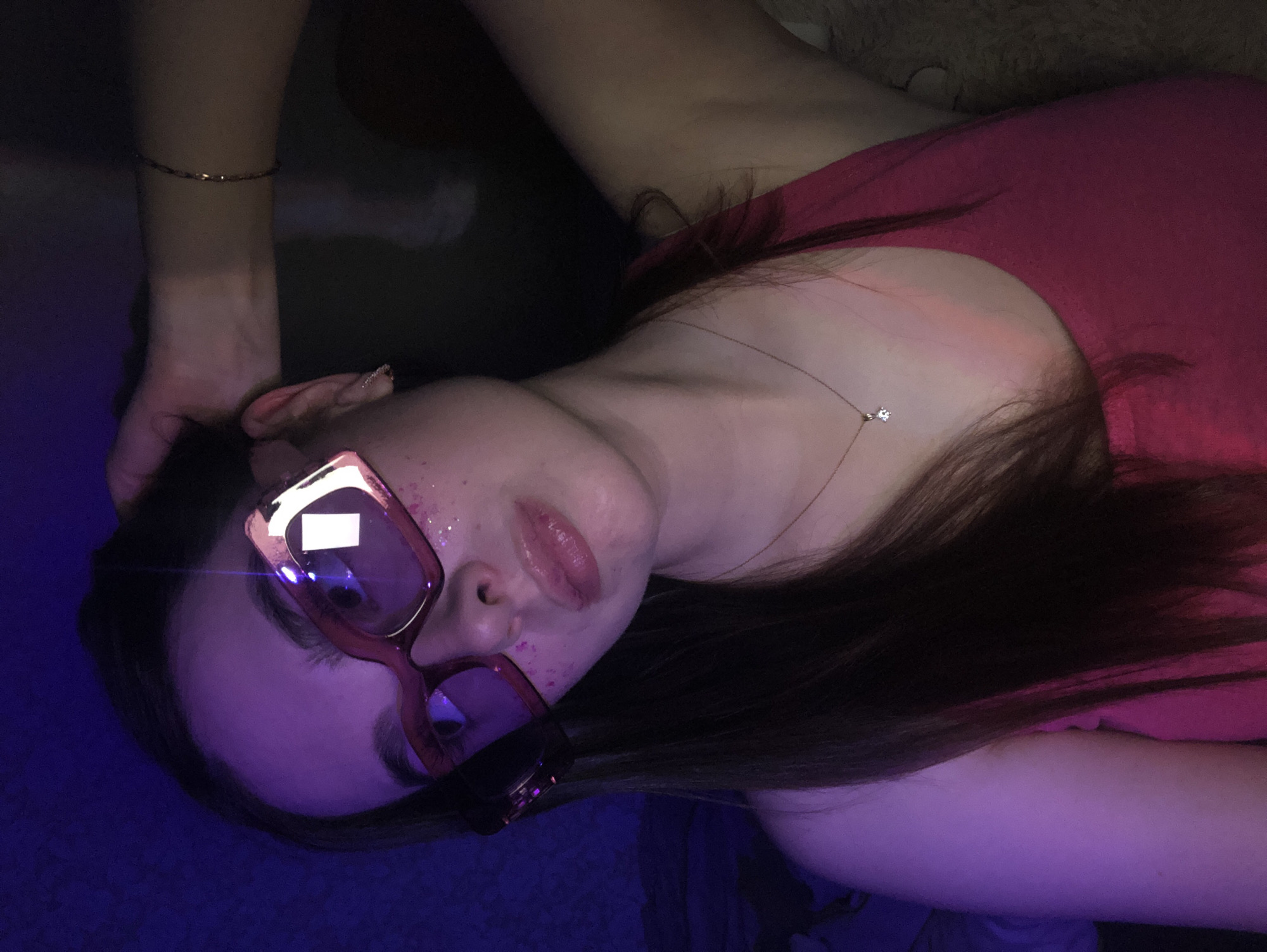 The image features a person, likely female, lying down indoors in what appears to be a dimly lit room in Noviy, Russia. The subject is wearing a vibrant pink top or dress and has long, dark hair that flows around them. Their head is tilted, and they are looking towards the camera.
The person is wearing distinct pink-framed sunglasses, which reflect bright white light sources. Pink glitter is visible on their face, specifically on the cheek and around the nose area, adding a decorative element. Their lips are slightly parted and appear to have a glossy finish.
Accessories include a delicate necklace with a small, sparkling pendant, visible on their chest, and a thin bracelet on their right wrist. The lighting is a notable detail, characterized by a dominant purple and blue hue illuminating the left side of the frame and parts of the subject's face, creating a cool and atmospheric ambiance. This contrasts with warmer, reddish-pink tones on the subject's upper body and hair on the right side. The background is dark and indistinct, suggesting a relaxed, low-light environment. The subject's pose is casual and relaxed, with their right arm extended above their head. No discernible text is visible in the image.