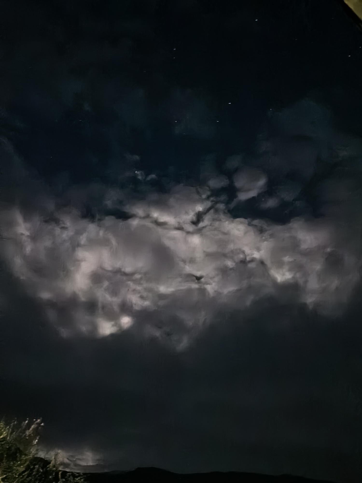 The image shows a night scene of clouds and stars over a silhouette of what appears to be a hill or mountain range. The sky is dark, with a band of illuminated clouds stretching across the middle of the frame. The stars are visible in the darker upper portion of the sky. In the lower left, there is vegetation, possibly grass or shrubs, that is partially illuminated. The exact location is unknown.