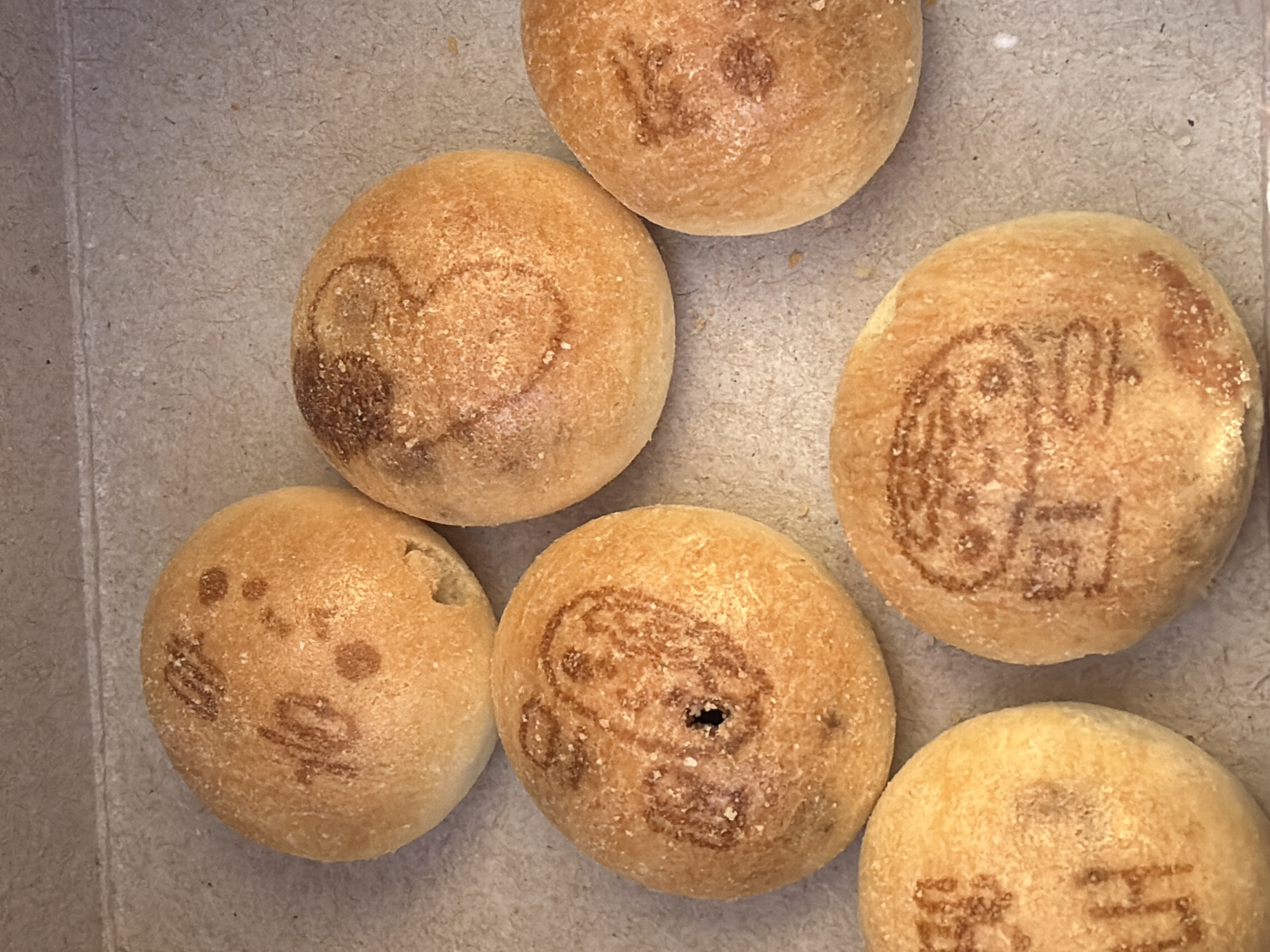 The image displays six roughly spherical, golden-brown baked items, likely buns or small loaves, arranged on a flat, light brown, textured surface consistent with recycled paperboard, possibly inside a box. Each item is dusted with fine white particulate matter. Five of the six baked goods exhibit distinct dark brown, baked-on or stamped patterns on their superior surfaces. These patterns include abstract shapes, a design resembling a simplified character or face, and another with partially discernible Cyrillic-like symbols. One item, featuring a character design, also presents a small, dark, circular indentation on its top surface. No individuals or actions are depicted. This scene is observed in Veliko Tarnovo, Bulgaria.