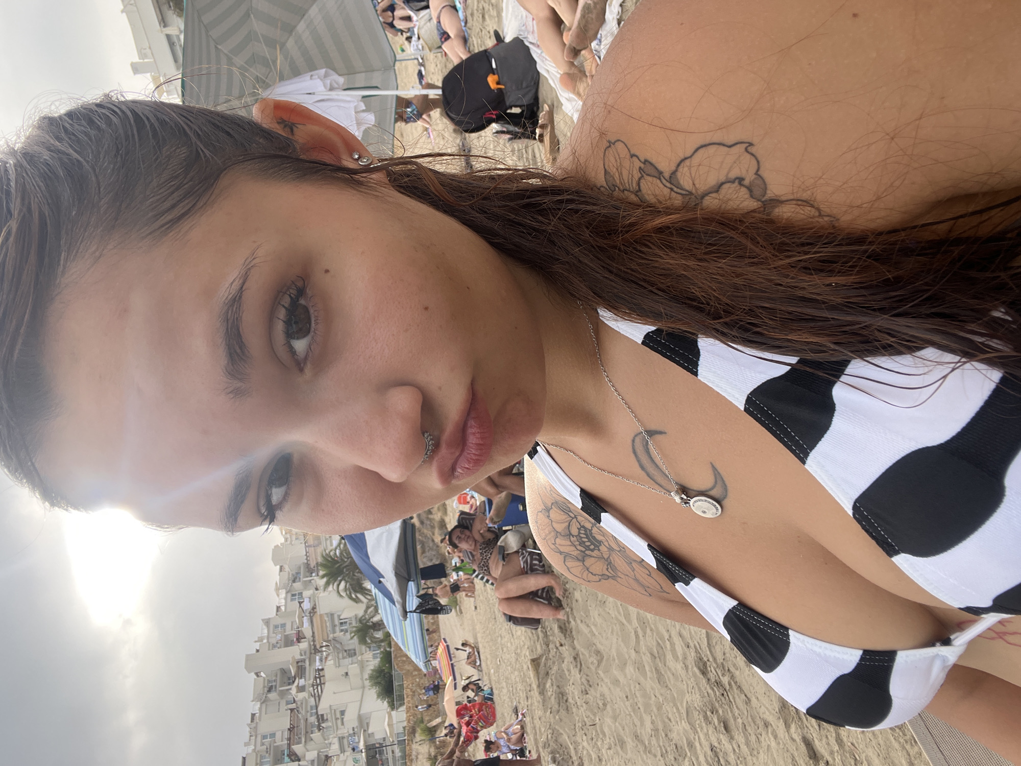 An outdoor, close-up, eye-level selfie shows a woman with wet, dark brown hair and a septum piercing at a beach in Urbanització Venta De Lanussa, Spain. She is wearing a black and white cow-print bikini top and a silver necklace with a crescent moon pendant. The woman is making a puckered-lip expression. In the background, the sandy beach is populated with other beachgoers, umbrellas, lounge chairs, and buildings. The sky is overcast with the sun peeking through.