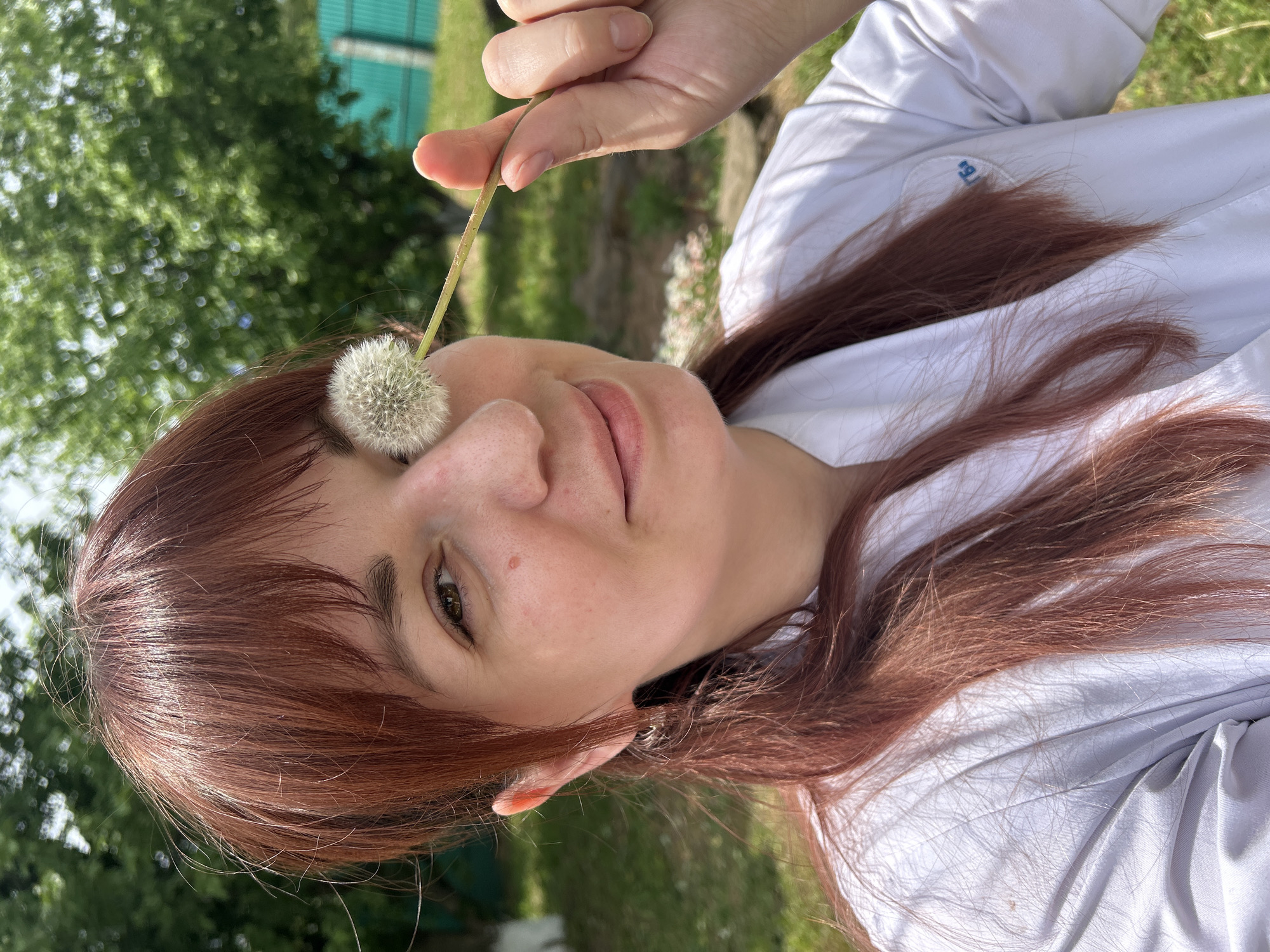 A woman is holding a dandelion in front of her face in an outdoor setting. She is wearing a white shirt and has reddish-brown hair. The background includes green trees and a green fence. The location cannot be determined to be in Фоки, Russia.