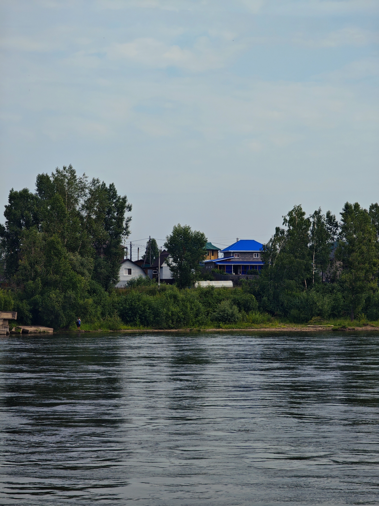 The image shows a wide river in the foreground, with its surface rippled by a gentle current. On the opposite bank, a dense line of trees forms a lush green backdrop. Scattered among the trees are several houses, varying in style and color. One house stands out with a bright blue roof. Power lines are visible strung between poles, suggesting the presence of electricity. On the left side of the riverbank, a concrete structure, possibly a pier or embankment, is partially submerged. Further to the right along the bank, a lone figure, appearing to be a person, stands near the water's edge, looking towards the river. The sky above is a pale blue, with wispy white clouds scattered across it. The overall scene suggests a peaceful, rural setting near a body of water.