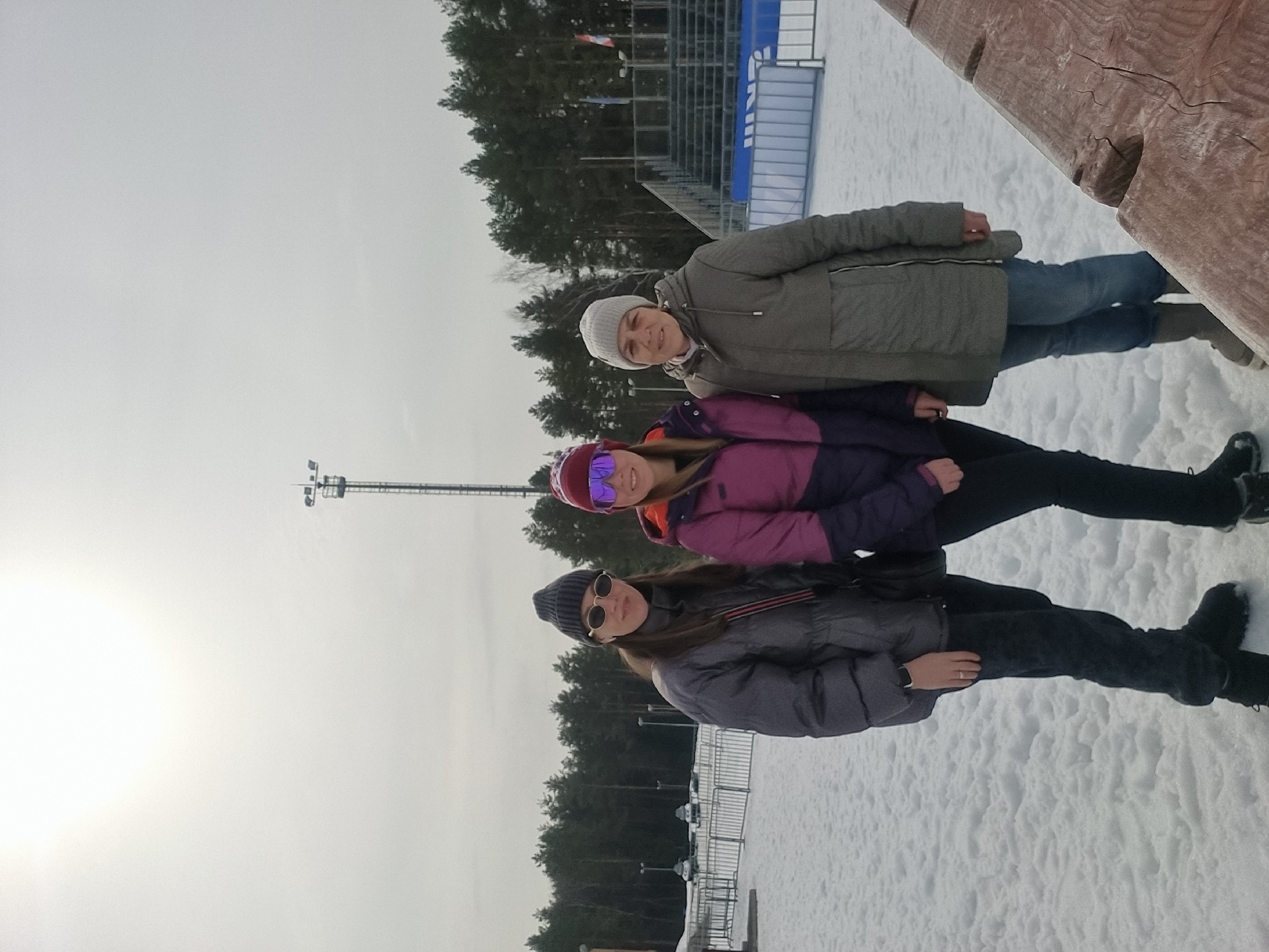 The scene depicts an outdoor winter environment at a sports venue, identified as being in Прикамский, Russia. The ground is covered uniformly with textured snow under a bright, overcast sky with a visible solar glare in the upper left. In the foreground, three adult females are standing, facing the camera. From left to right:
1. A female wearing a dark grey knitted beanie, small oval sunglasses with light frames, a dark grey quilted jacket, dark trousers, and dark winter boots. She carries a dark shoulder bag with a red and black striped strap, and a ring is visible on her left hand's fourth finger.
2. A female wearing a dark red knitted beanie, mirrored purple sports sunglasses, a two-tone purple and dark purple winter jacket with orange accents, dark trousers, and dark winter boots.
3. A female wearing a light grey knitted beanie, an olive-green parka, blue jeans, and light grey winter boots.
All individuals appear to be posing for the camera.
The background features a dense evergreen forest. Directly behind the individuals, a tall metal light tower with multiple floodlights is prominent. To the right, multi-tiered stadium bleachers or grandstands are visible, partially obscured by blue and white barrier fences. On the far left, a white metal barrier separates the foreground from a distant area containing several small vehicles and utility poles. A section of a rough-hewn wooden structure occupies the lower right corner of the image.