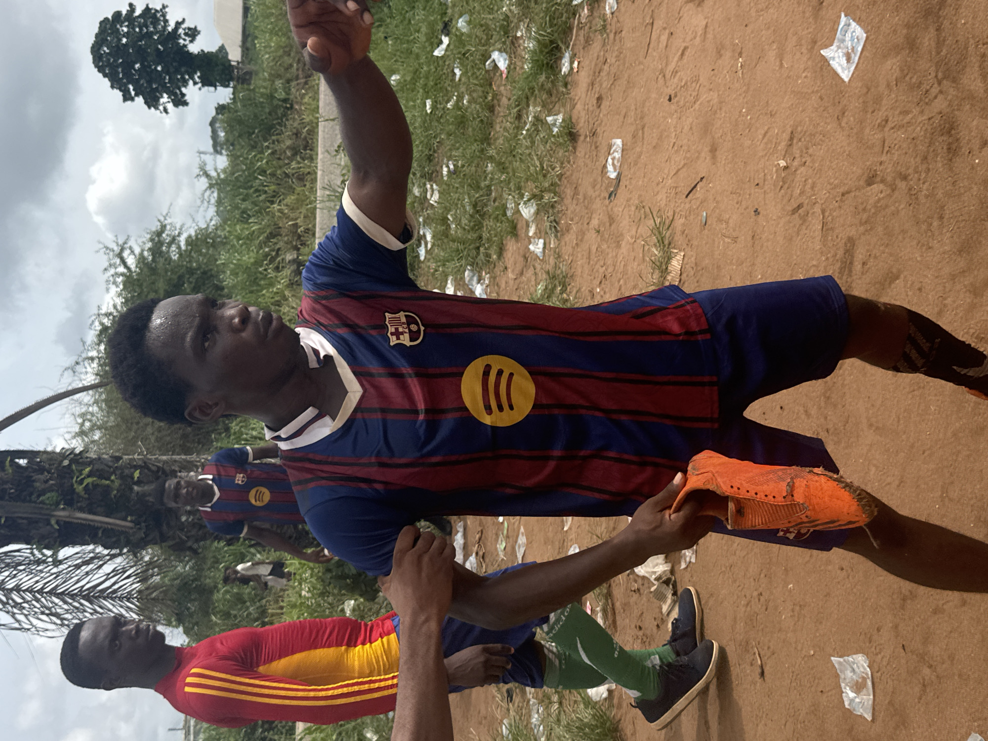 The image shows three men outdoors in Ihiagwa, Nigeria. The man in the foreground wears a blue and maroon FC Barcelona soccer jersey with a white collar and a yellow Spotify logo. He holds a worn orange cleat in his left hand. To the left, another man wears a red and yellow soccer jersey and green knee-high socks. In the background, a third man also wears the same Barcelona jersey. The ground is dirt and grass, littered with white pieces of trash. There is a tree in the background under a cloudy sky.