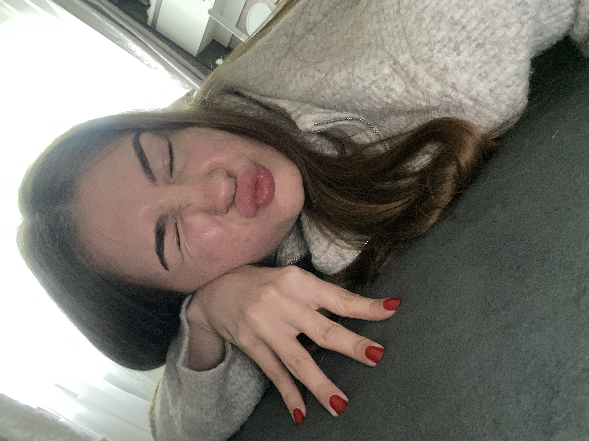 A young woman is featured in a close-up, diagonally oriented shot, appearing to be lying on her side. She has long, dark brown hair that falls around her head and shoulder. Her face is directed towards the camera, showcasing a playful and exaggerated expression with eyes squinted and lips tightly pursed into a "fish face" or "duck face." A silver hoop nose ring is visible on her left nostril.
She is wearing a light gray, possibly knitted or textured, long-sleeved top or jacket, indicated by a visible silver zipper on the shoulder. Her right hand, with fingers slightly splayed and nails painted bright red, rests on a dark green or dark gray fabric surface, likely a sofa or bed. The background is brightly lit and overexposed, suggesting a window or strong light source, with faint architectural details like curtains or a ceiling visible. The scene takes place indoors, likely during the daytime. There is no visible text in the image. The overall mood conveyed is casual and humorous.