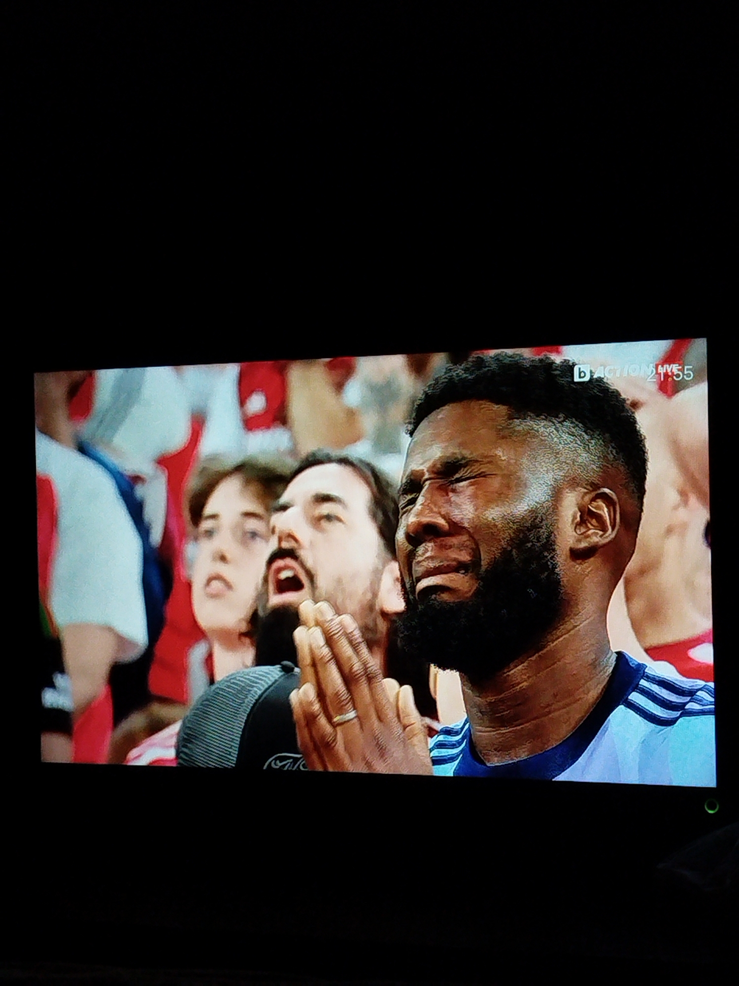 The image shows a close-up of three spectators at a sporting event. A Black male with a beard is crying with his eyes closed. To his left is a man with a beard and a woman, both looking up with anticipation. The man has his hands clasped together in a praying gesture. The spectators are wearing various clothing, including a light blue and white striped jersey. The background shows other spectators wearing red and white. There is a sports broadcast watermark in the upper right corner of the image. The location of the image is not discernible.
