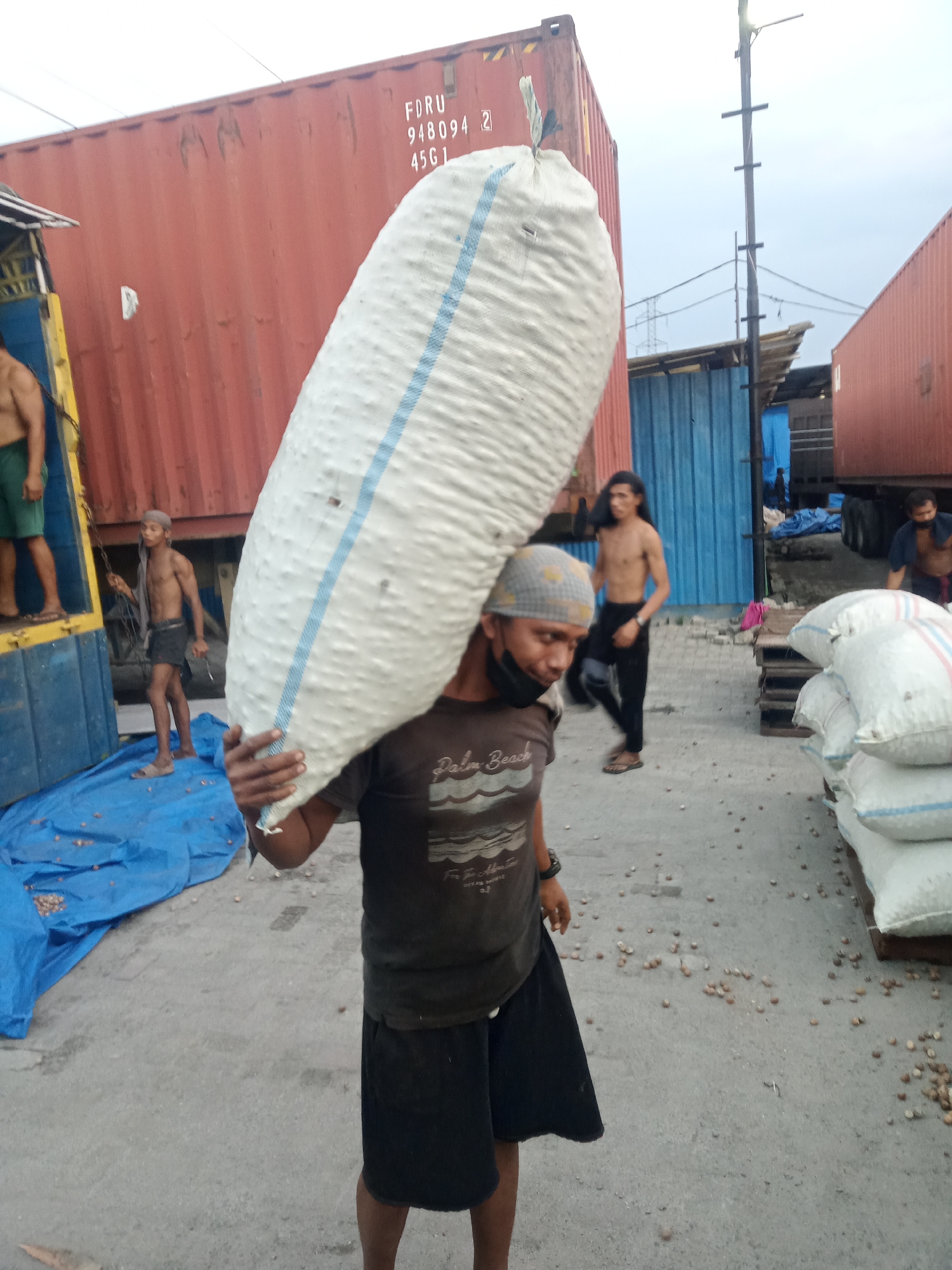 In Sunggal, Indonesia, a man is captured carrying a large, white sack across his shoulders. The sack, possibly filled with goods, is elongated and appears to be made of woven material with two thin blue stripes running vertically. The man wears a gray bandana, a dark t-shirt with a graphic print, and dark shorts. He is also wearing a black face mask. The background reveals a gritty, outdoor setting with several large, red shipping containers, one of which has markings indicating it's a "FDRU 948094 2 4561". Another shipping container, painted blue, is visible to the left. The ground is paved and scattered with small, light-brown objects, possibly nuts or seeds. Several other individuals are present in the background, including one man partially visible in the left of the frame, wearing green shorts, and another man with long dark hair walking in the middle ground. Piles of white sacks are stacked on wooden pallets to the right of the frame. The lighting suggests it is daytime, with an overcast sky.