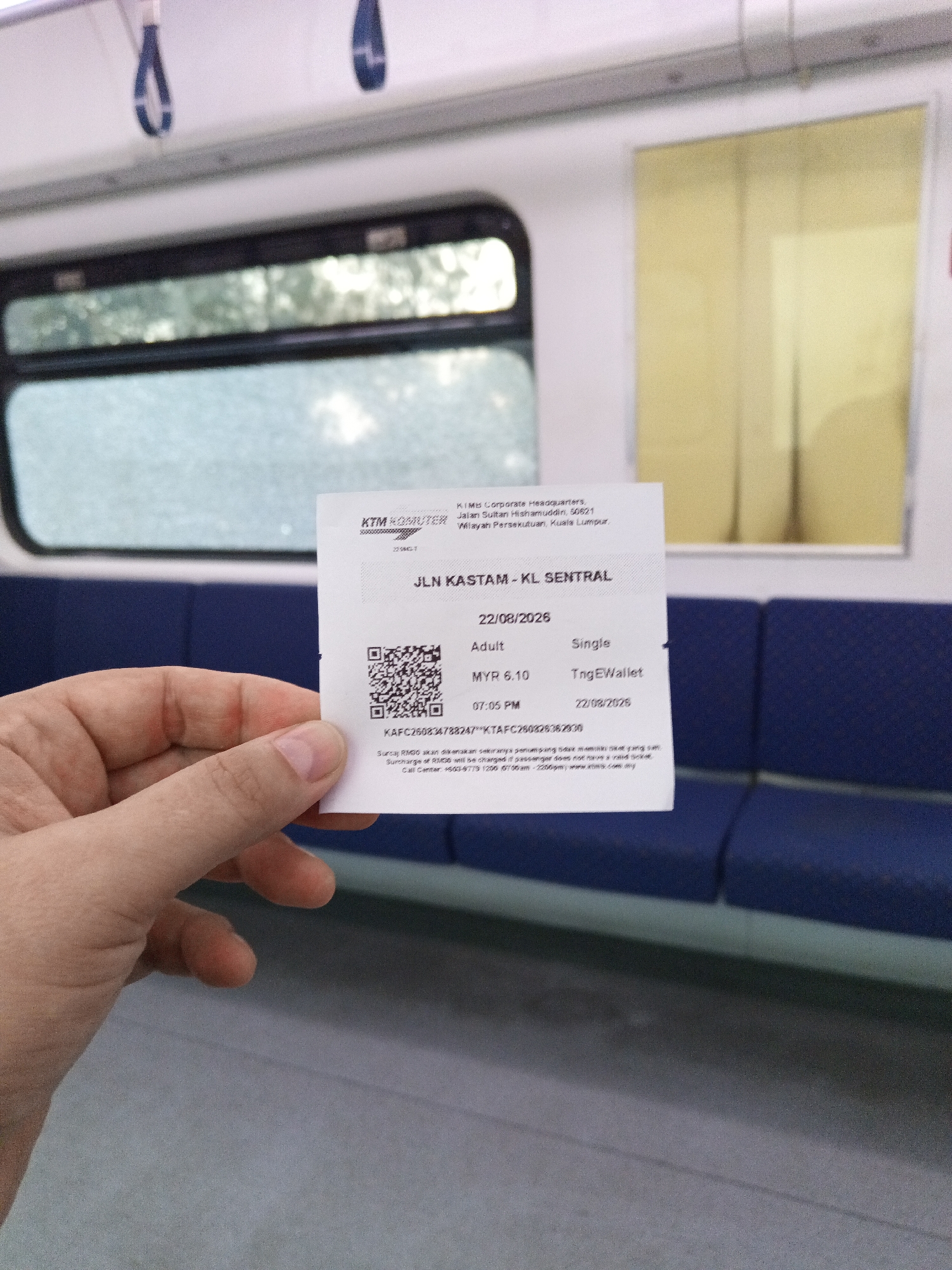 A close-up, slightly angled shot shows a person's hand holding a train ticket for KTM Komuter in Malaysia. The ticket is white with black text and a QR code. The ticket reads "JLN. KASTAM - KL SENTRAL", indicating the origin and destination. The date on the ticket is "22/08/2026" and the time is "07:05 PM". The fare is "MYR 6.10" and it is for an "Adult" ticket using "TngEWallet". The background of the image is the interior of a train carriage, with blue seats and windows. One window shows a blurred view of trees and sky. The overall environment suggests the person is traveling by train within Klang City, Malaysia.