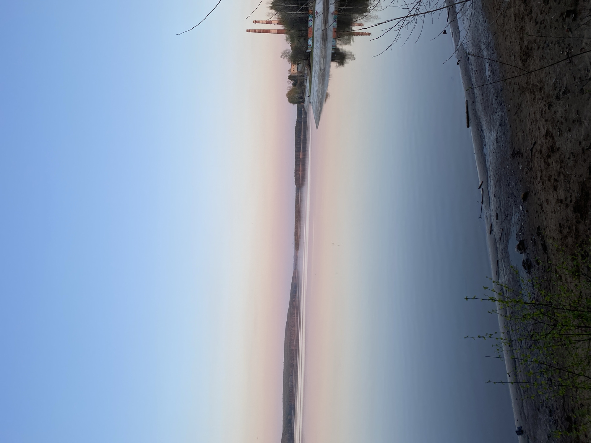 The image shows a landscape with a body of water, likely a river or lake, with a bank on the right side covered in sand and vegetation. The water reflects the sky, which has a gradient from blue to pink, indicating sunrise or sunset.
Across the water, on the opposite bank, there are buildings and industrial structures, including tall chimneys or towers. The buildings are reflected in the water as well. The image appears to be taken near Chaikovsky, Russia, based on the location of the industrial structures.